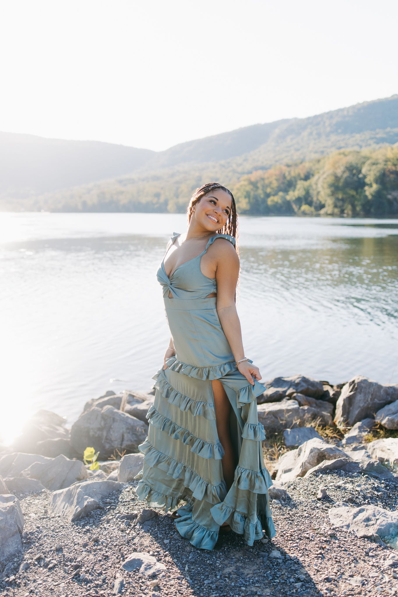 Woman in sage green ruffled maxi dress standing on rocky shore with mountain lake backdrop.