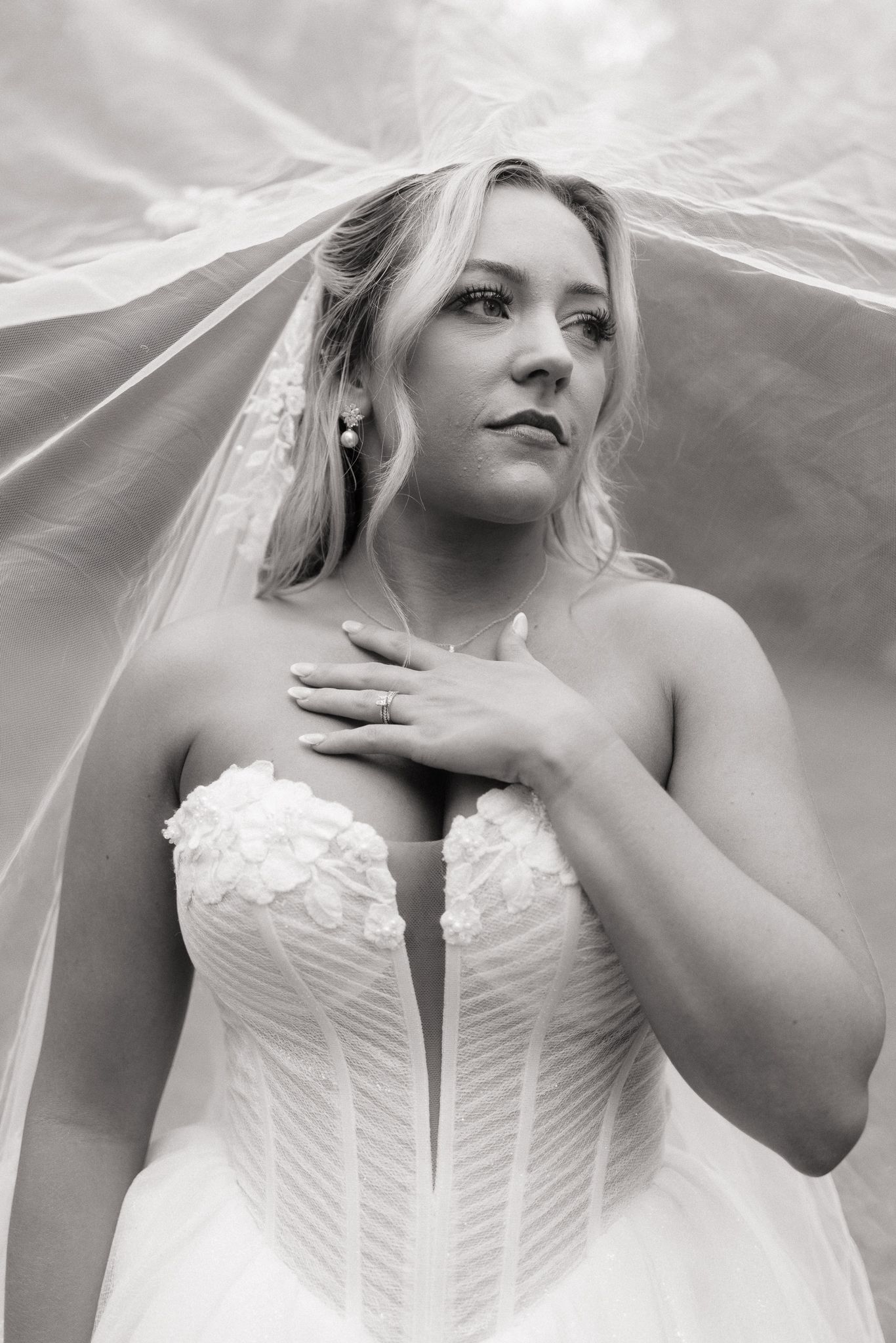 Black and white bridal portrait of a blonde bride in a strapless gown under a flowing veil, hand on chest.