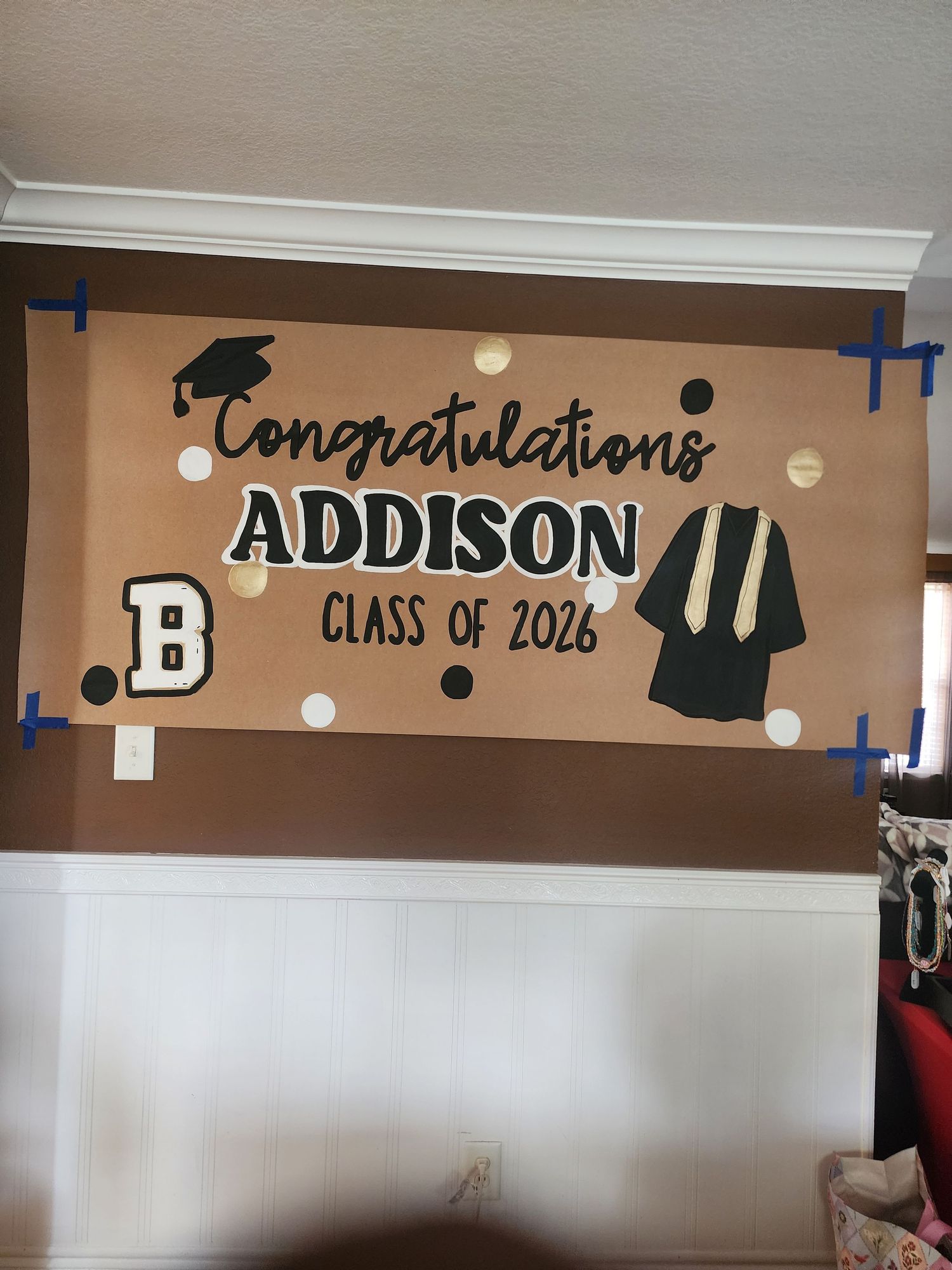 Handmade graduation banner congratulating Addison, Class of 2026, with gold and black decorations taped to a white wall.