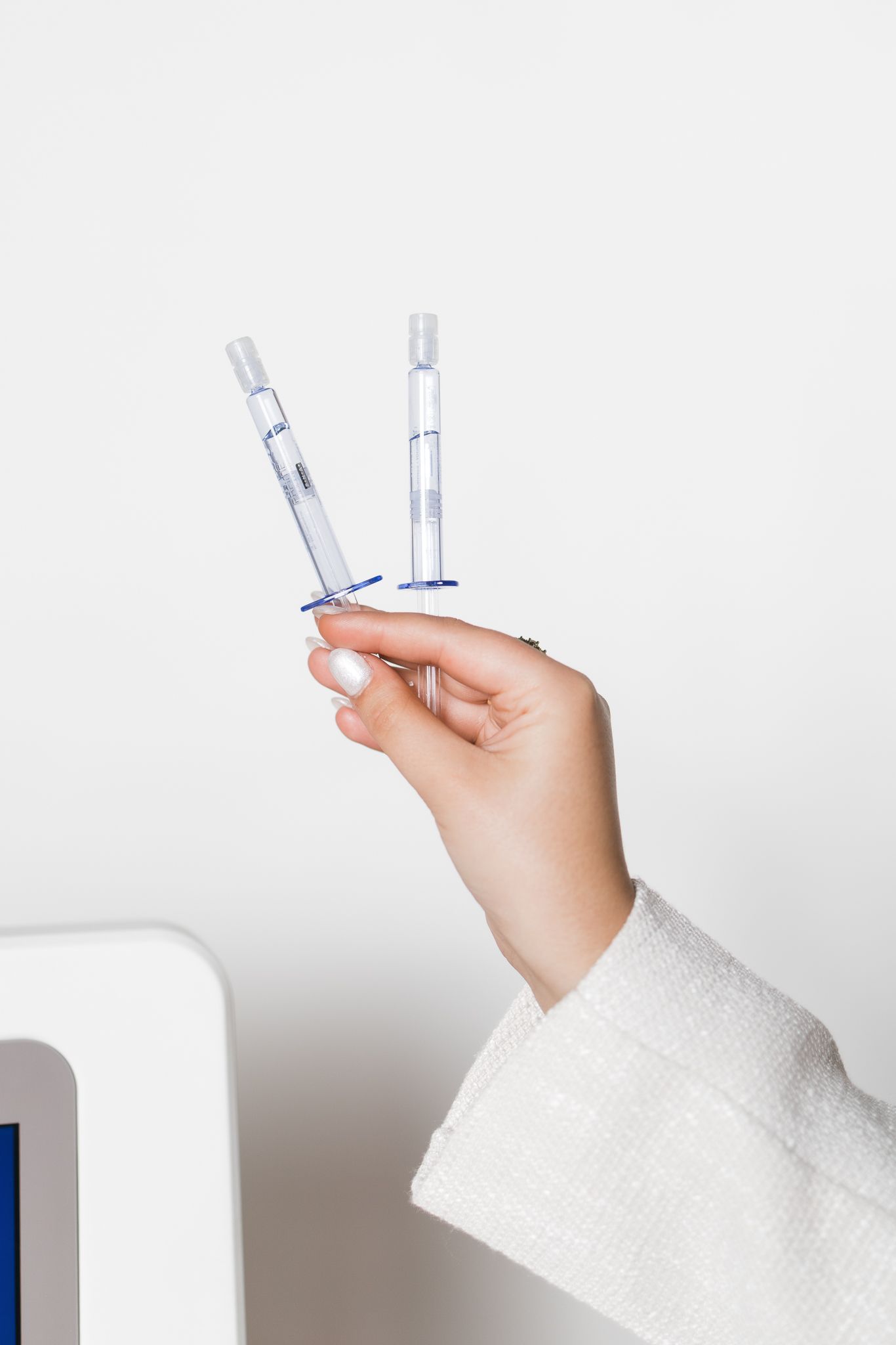Hand holding two clear medical syringes against white background near skincare device.