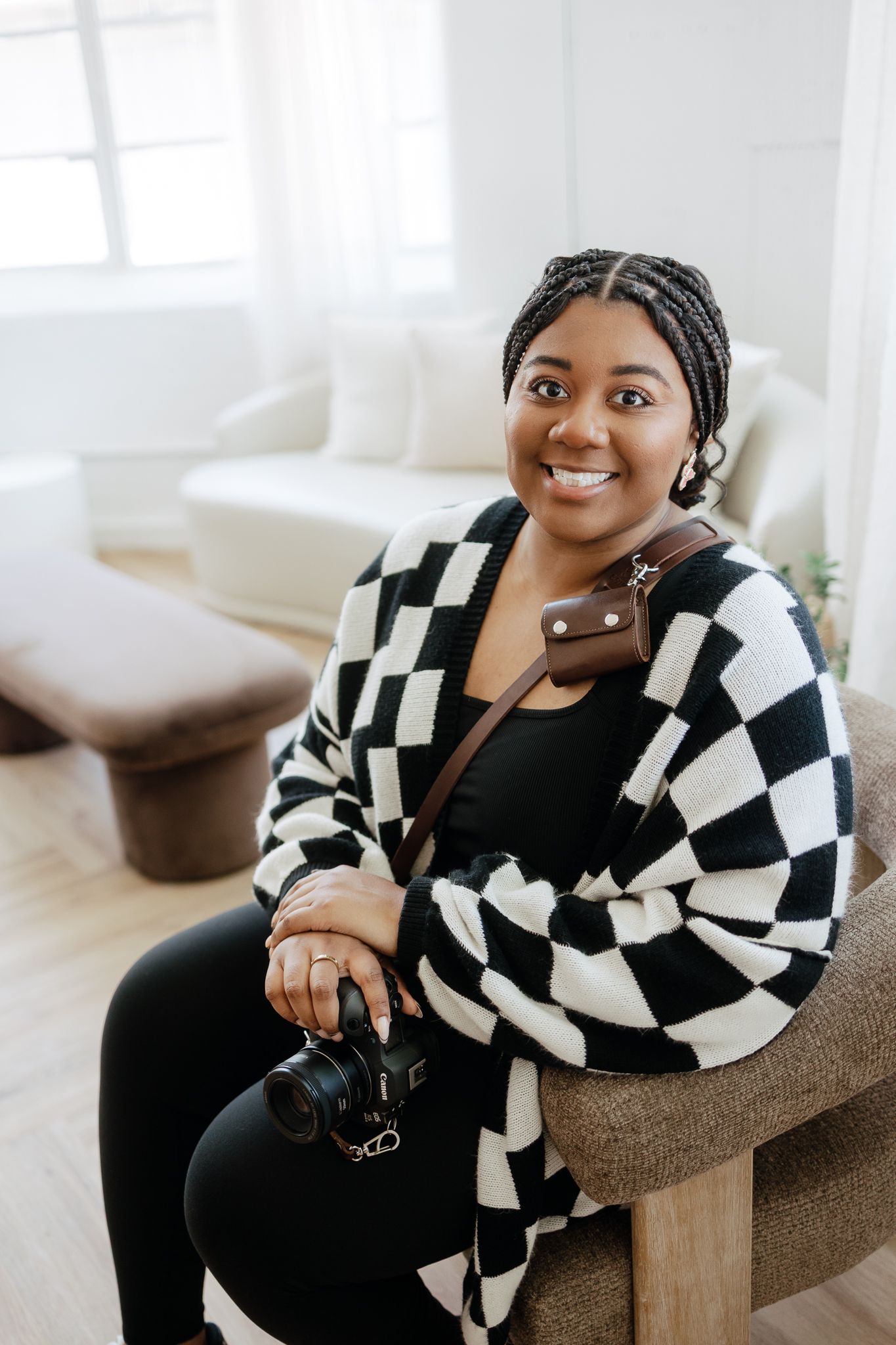 Smiling Black woman photographer sitting in chair holding camera wearing black and white checkered cardigan.