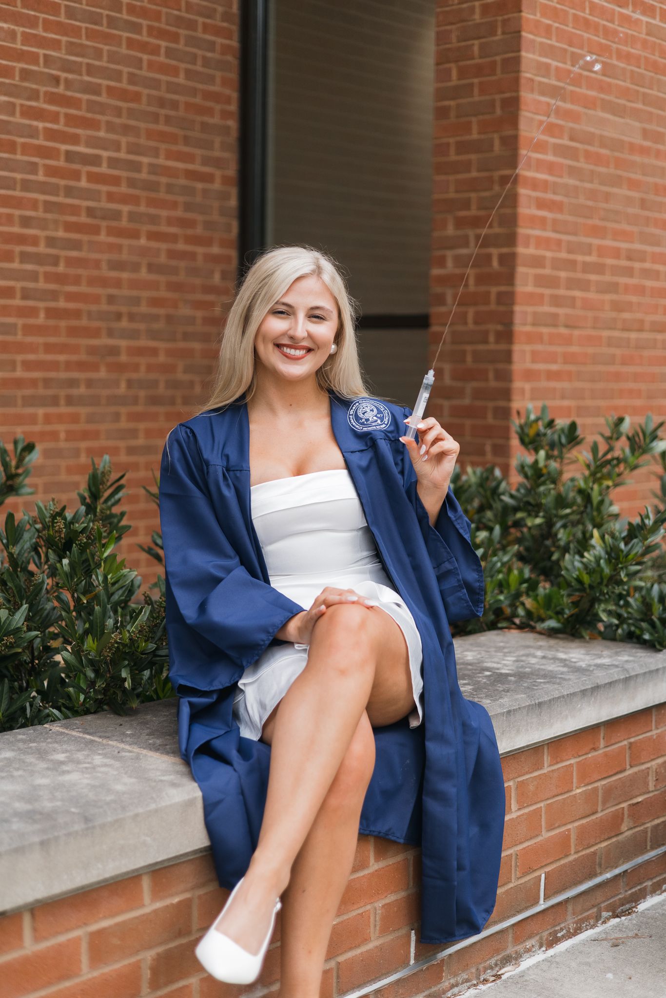 Smiling blonde woman in blue graduation gown and white dress sits on brick ledge holding diploma outdoors.