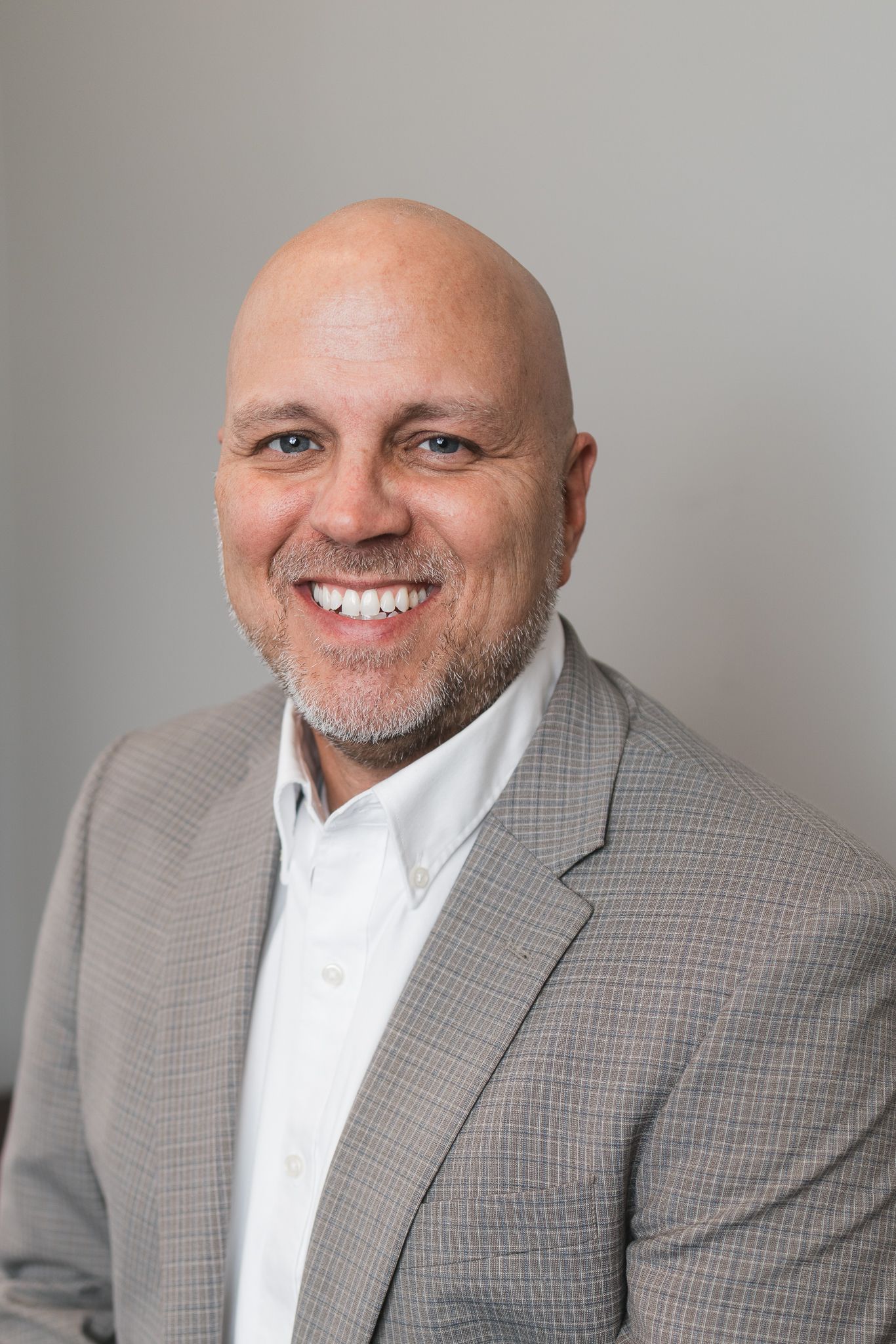 Smiling bald man with beard wearing a gray blazer and white shirt against gray background.