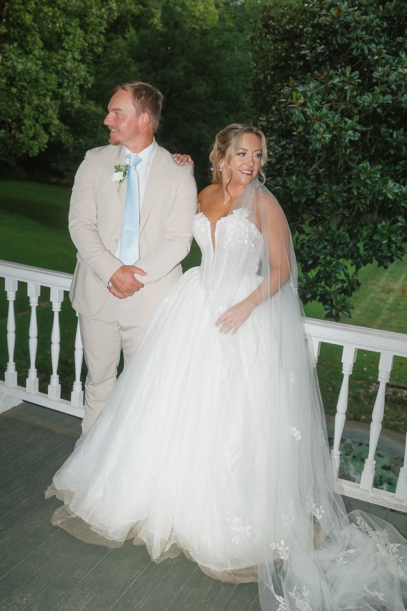 Bride in white ballgown and groom in tan suit pose together on a white porch railing surrounded by lush greenery.