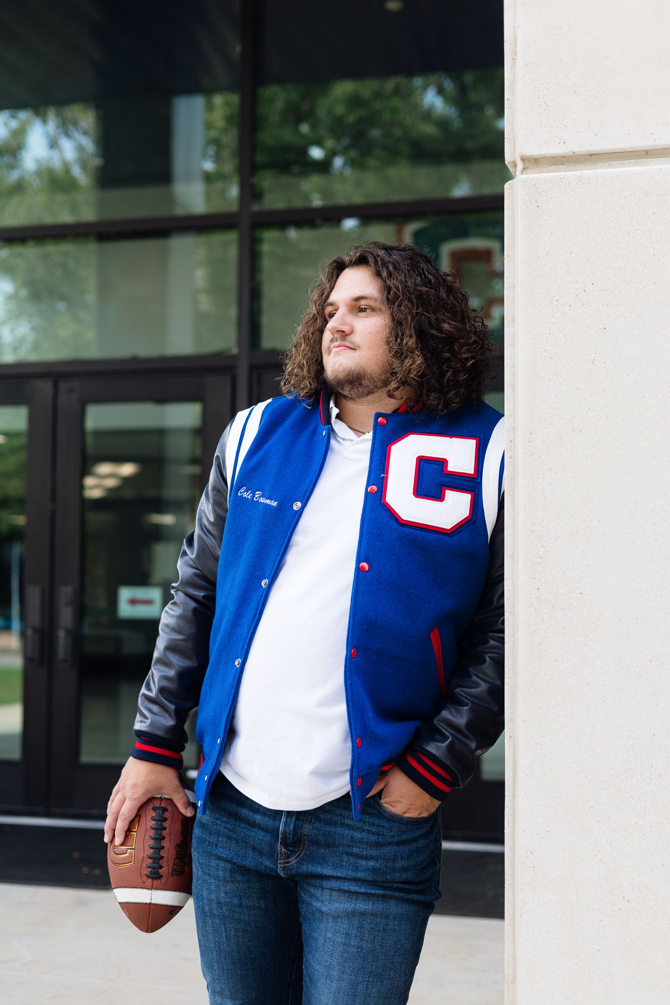 Young man with curly hair in blue varsity jacket holding football, leaning against column outside school building.