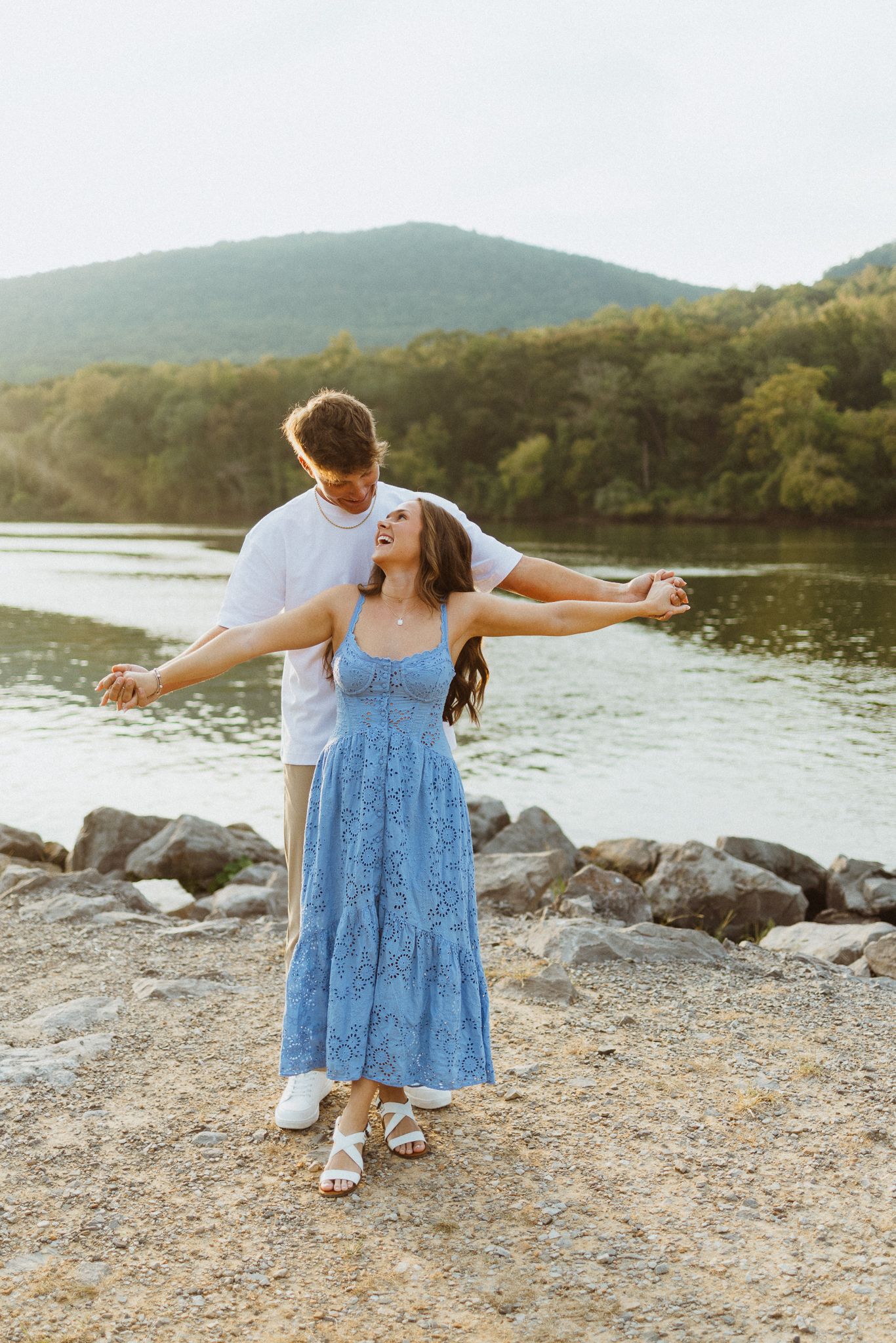 Couple dancing by rocky lakeside with mountain backdrop at golden hour sunset.