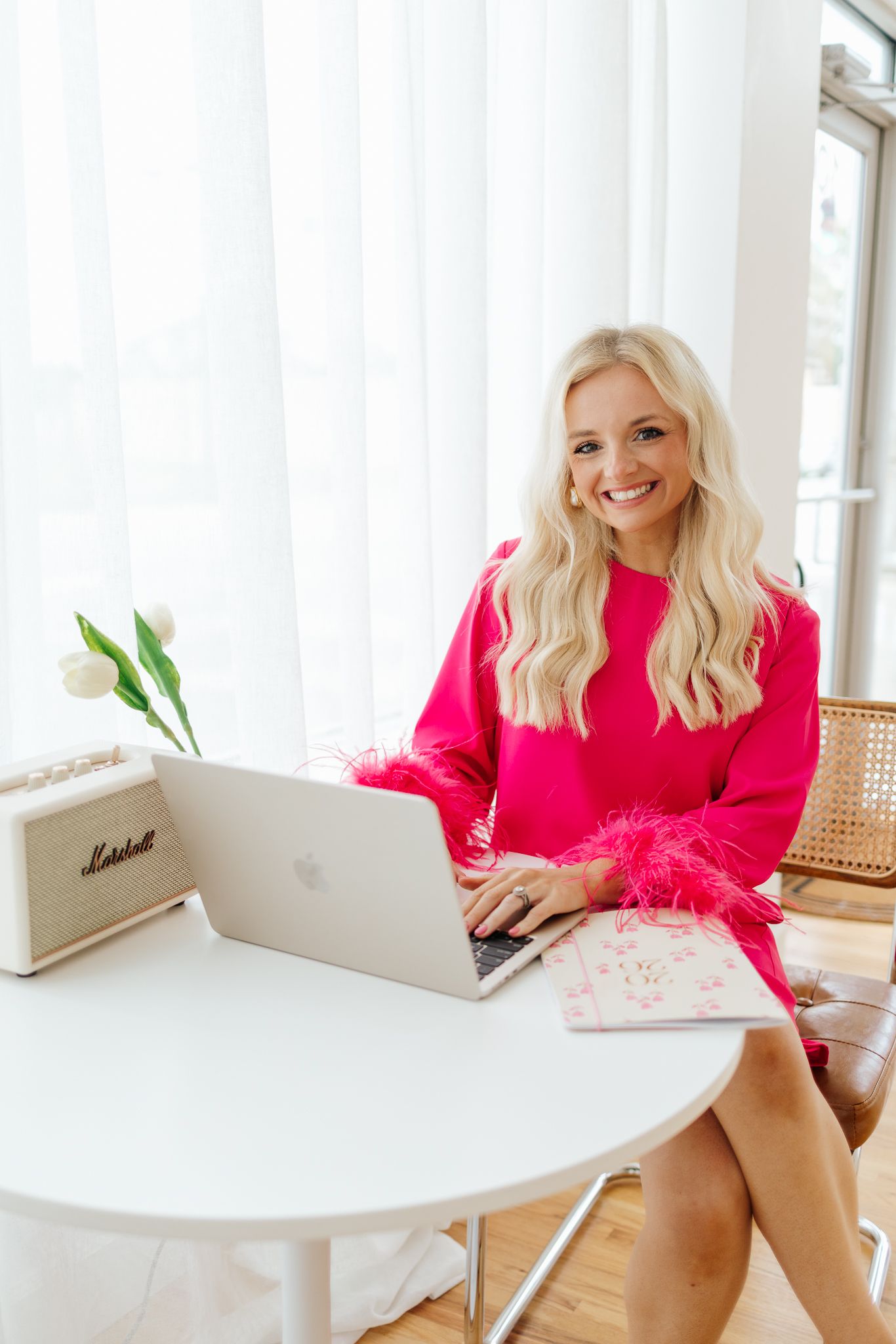Blonde woman in pink feather dress works on MacBook at white desk by bright windows.