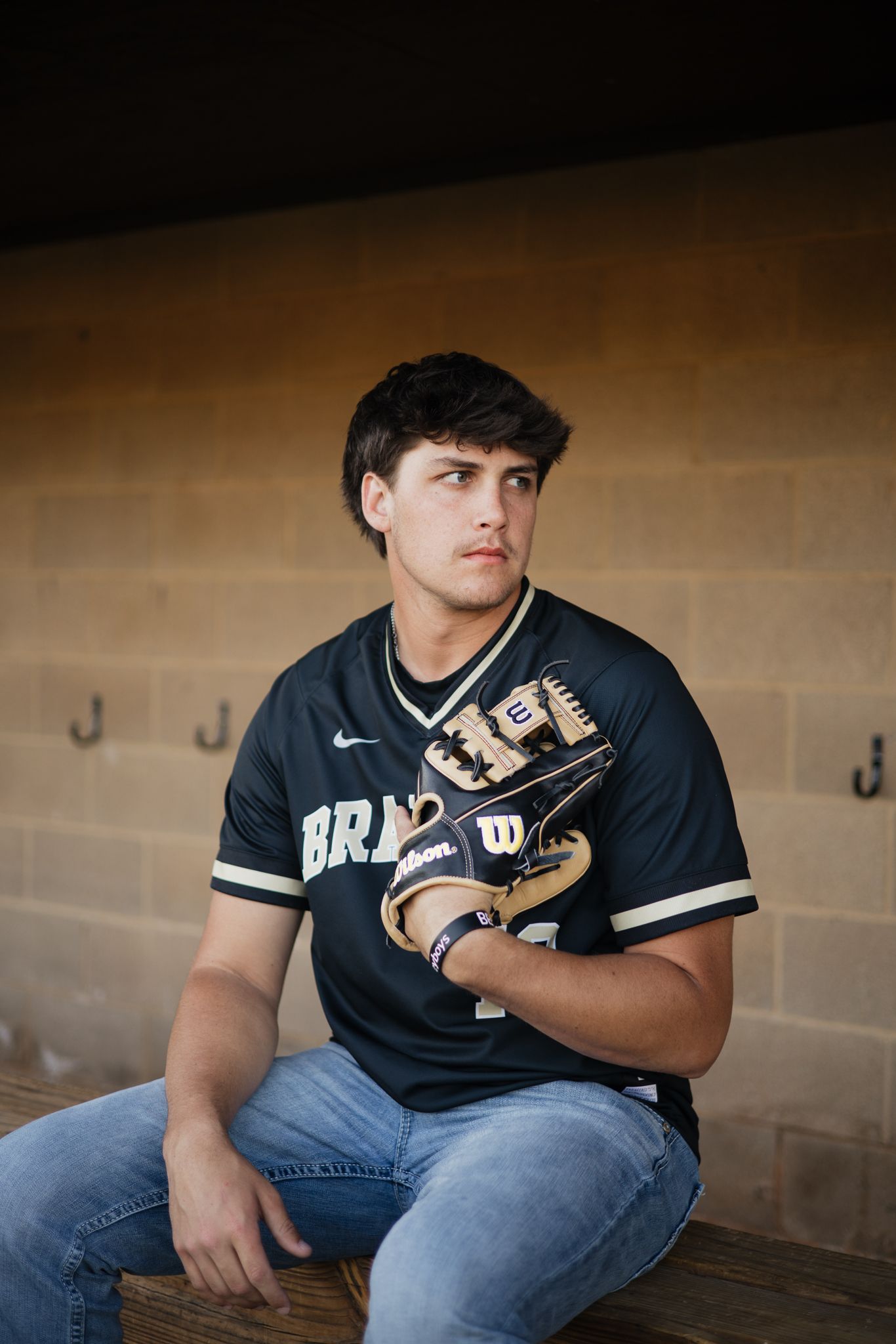 Young baseball player in a black Nike jersey holding a Wilson glove, sitting in a dugout wearing jeans.