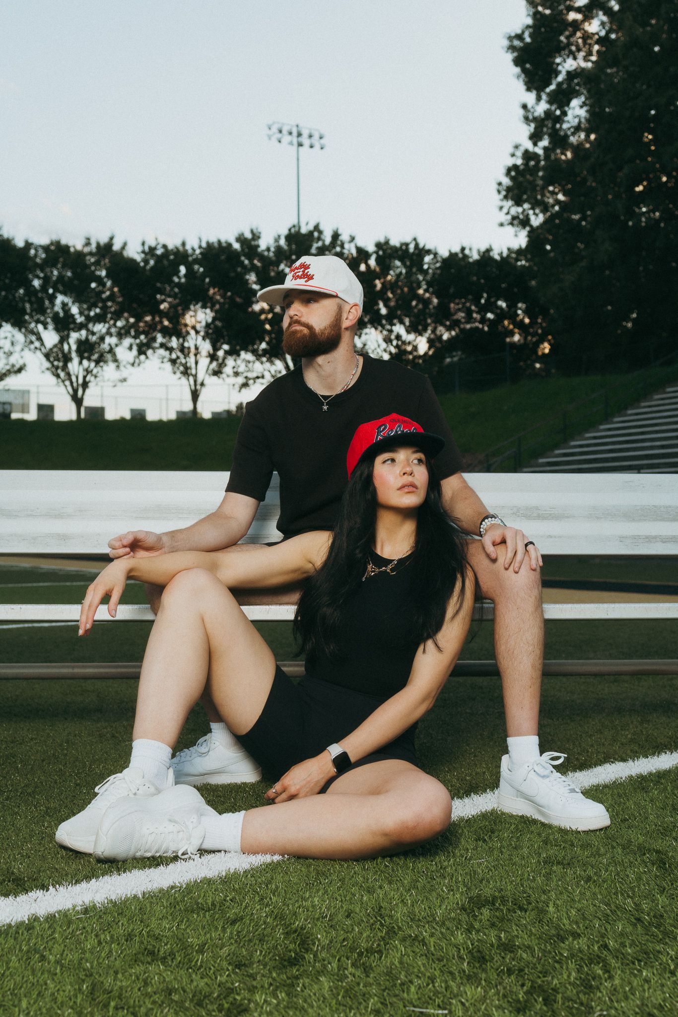 Man and woman sitting on bleachers wearing red and white baseball caps and black outfits.