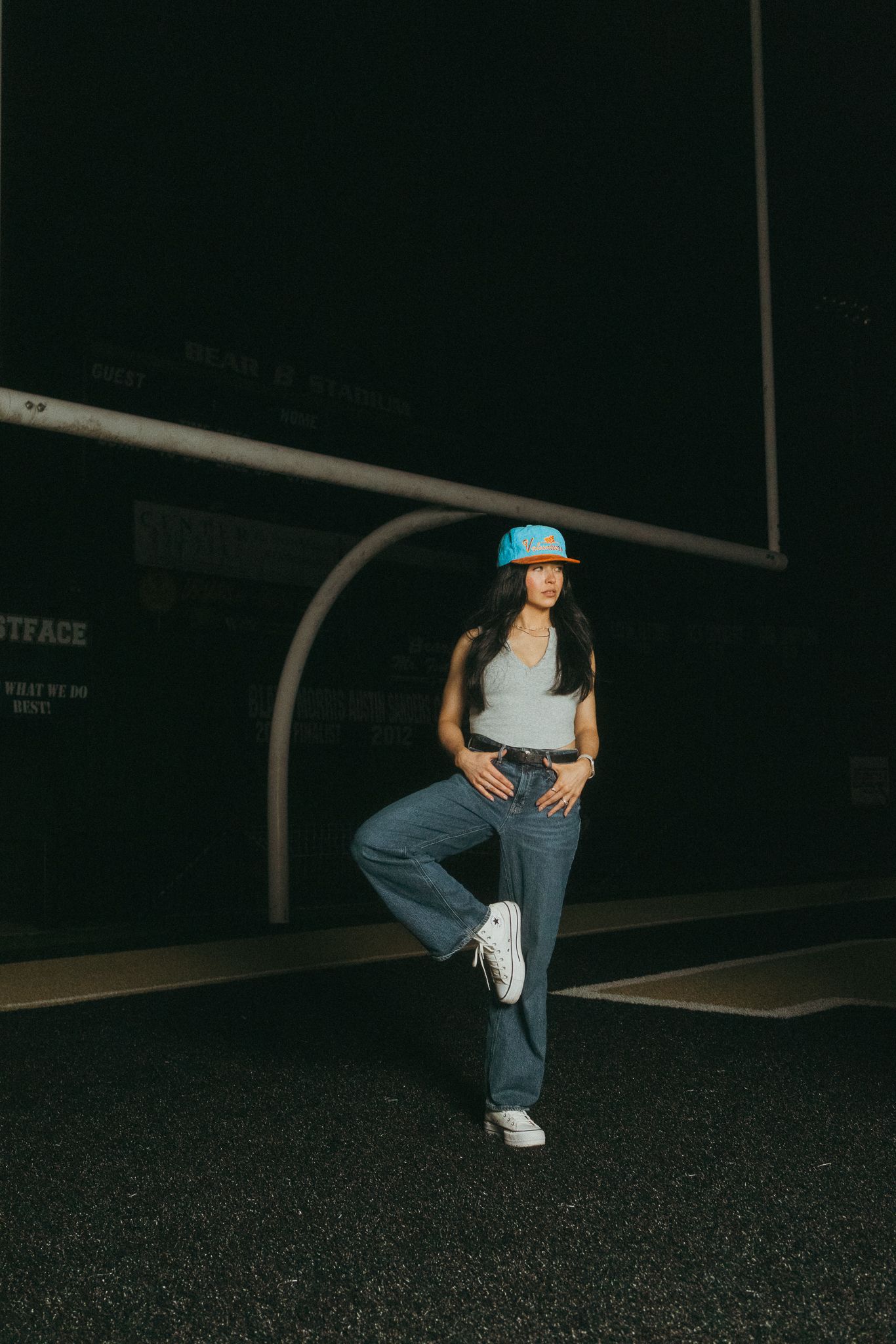 Young woman in blue bucket hat, gray tank top, and wide-leg jeans posing against dark urban background at night.