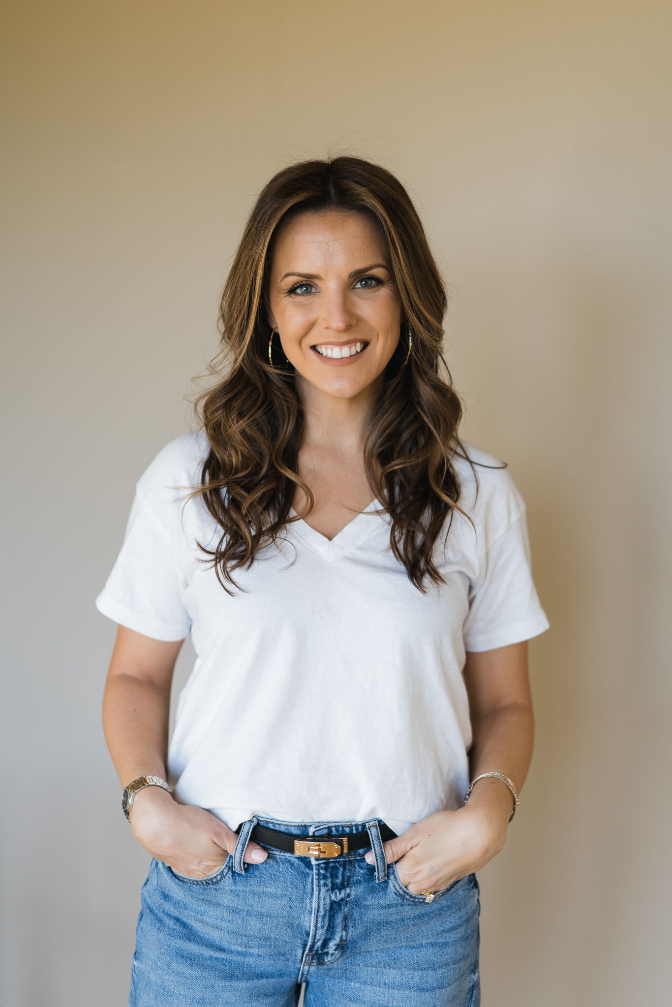 Smiling brunette woman in white v-neck tee and jeans posing casually with hands in pockets.