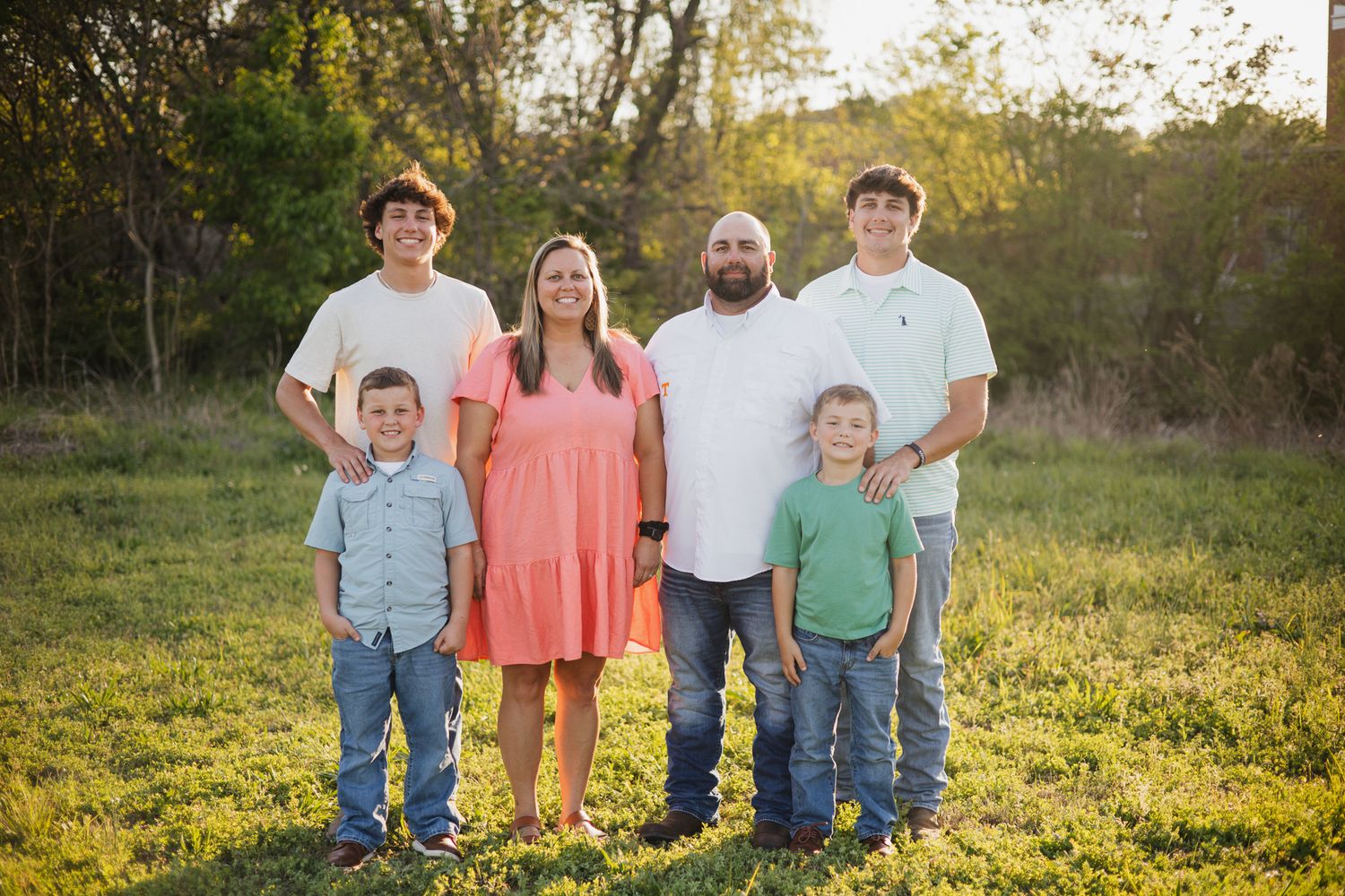 Family of six posing outdoors in a green field during golden hour, wearing casual spring attire.