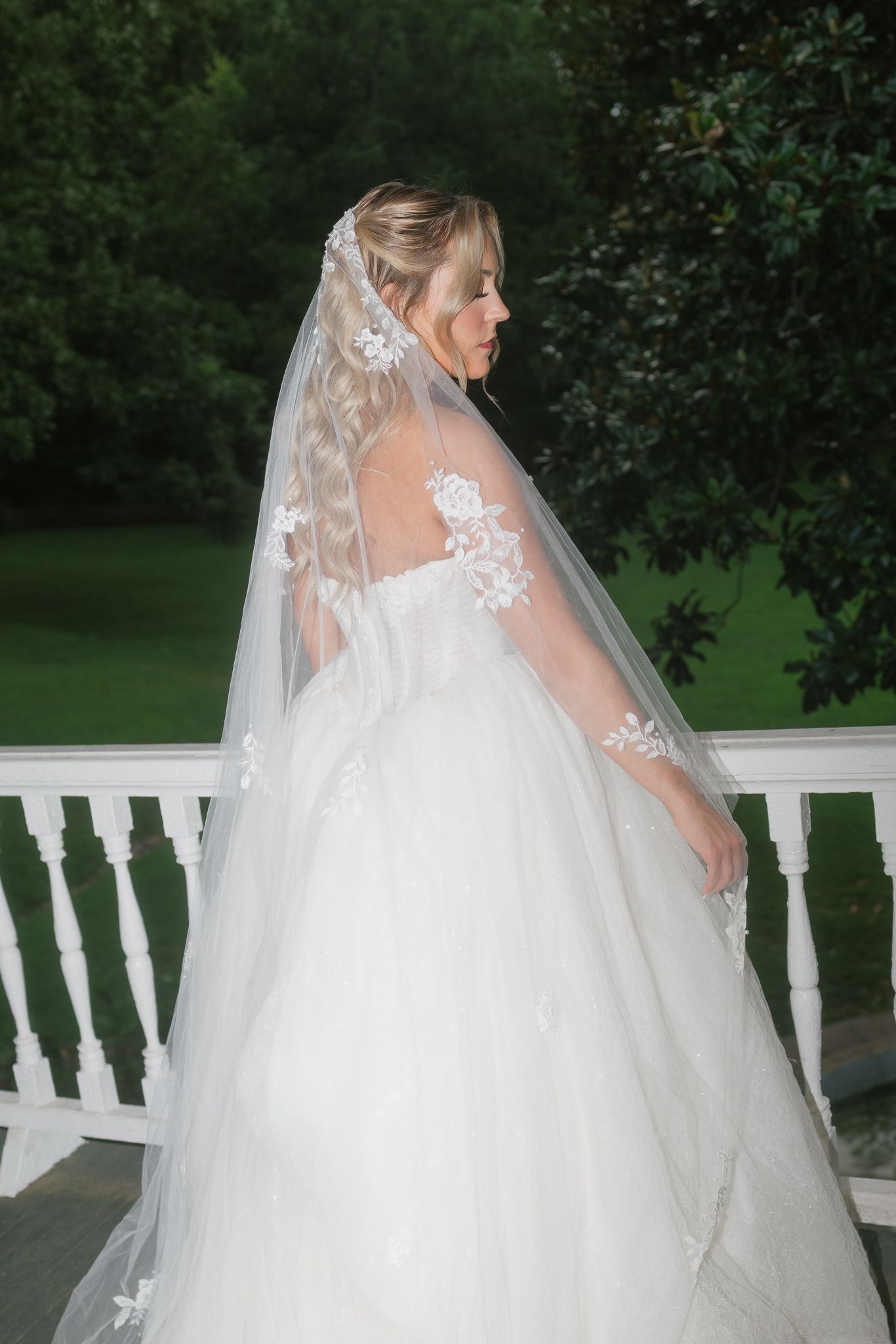 Bride in white ballgown with lace veil stands on porch surrounded by lush green trees.