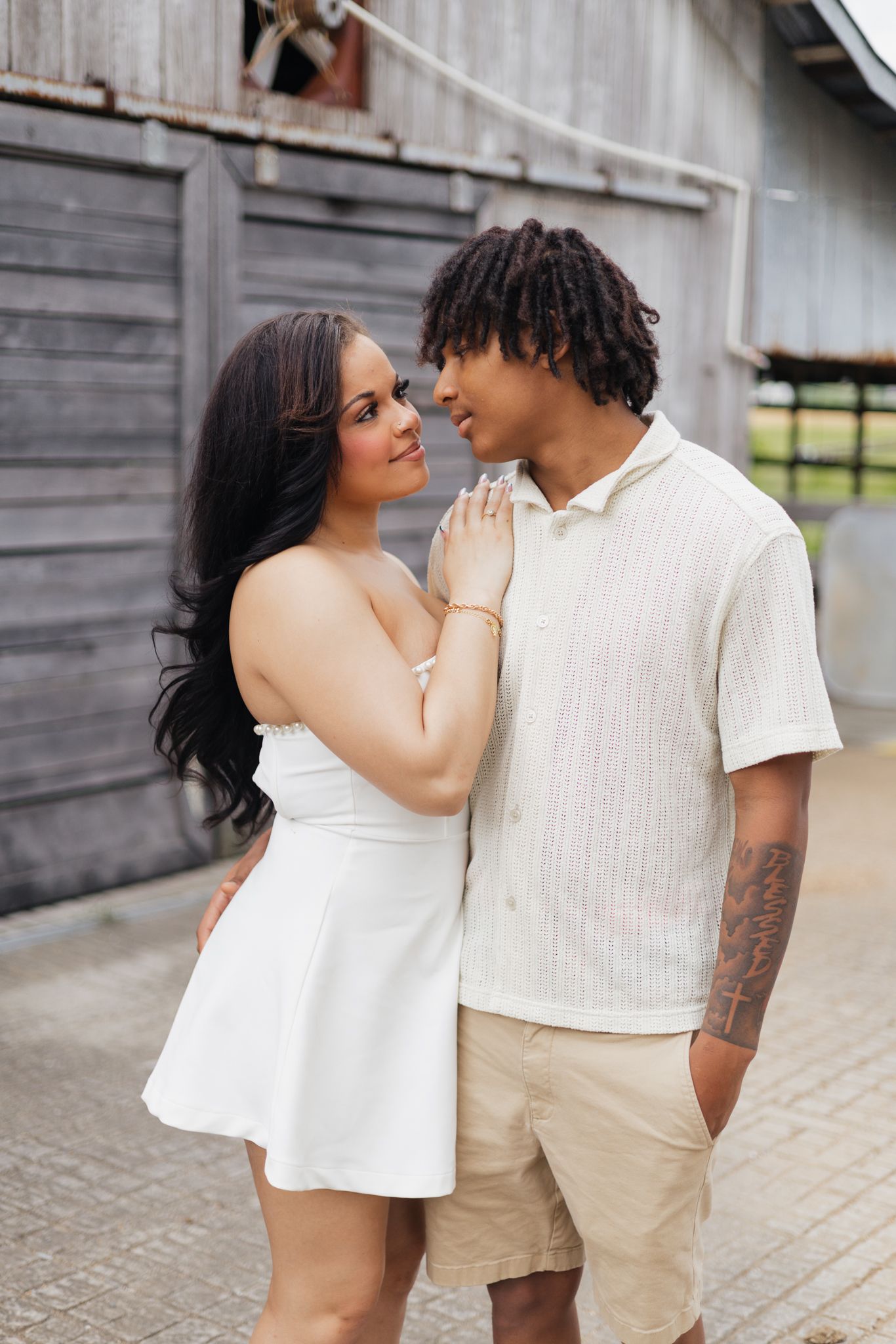 A young interracial couple in white outfits share a romantic moment outside a rustic barn.