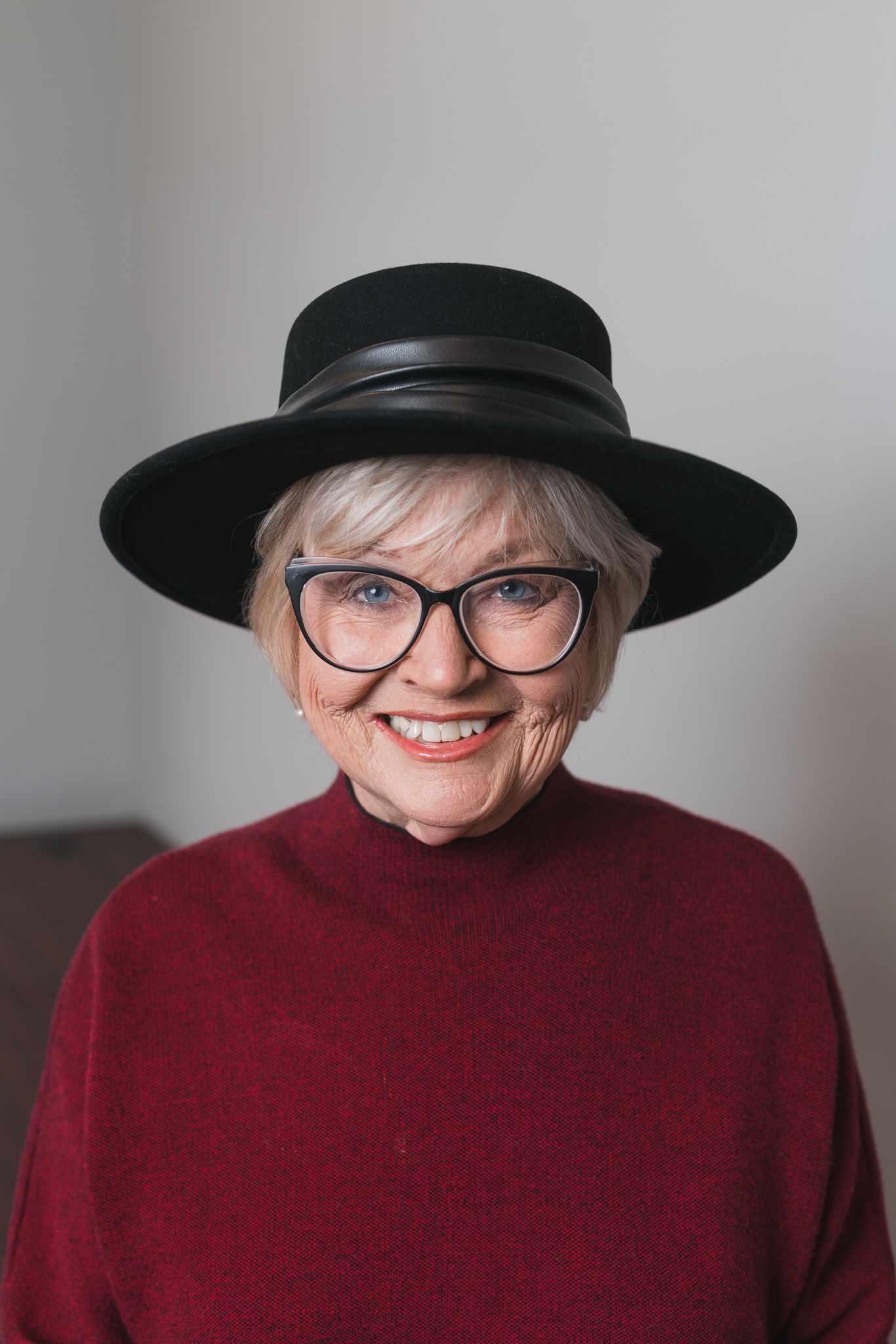 Elderly woman with short gray hair wearing black cat-eye glasses, a black hat, and red turtleneck.
