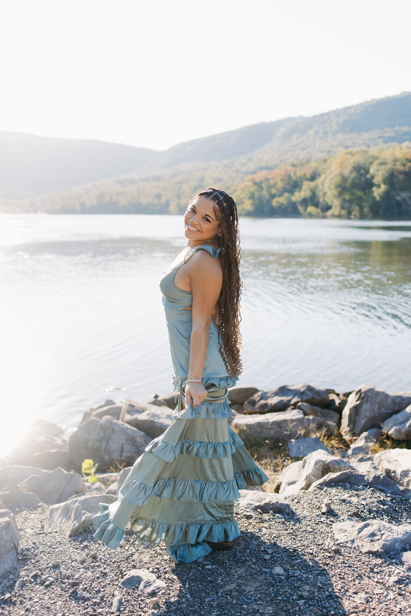 Young woman in a tiered blue dress posing by a rocky lakeside with tree-covered mountains in the background.