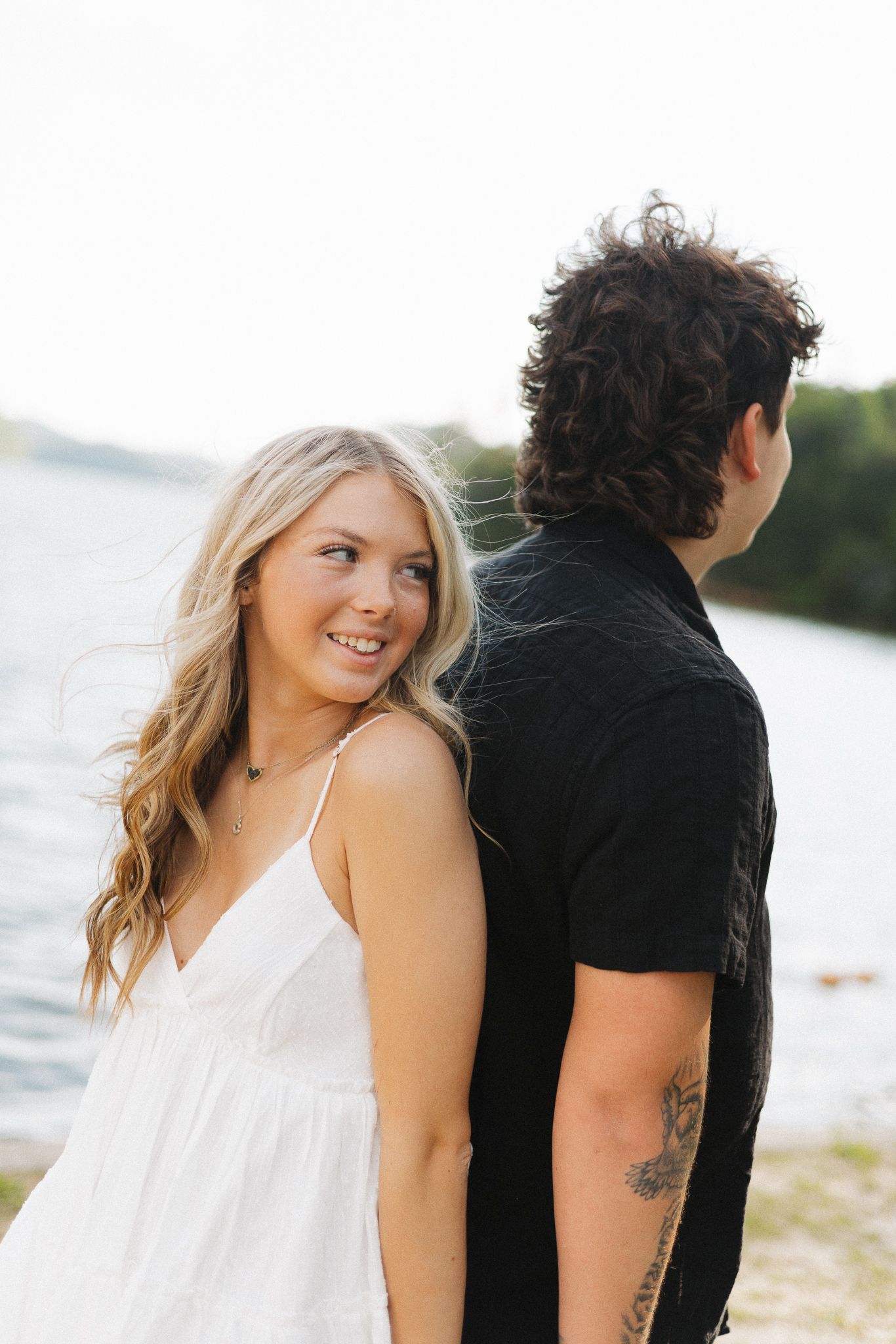 A smiling blonde woman in a white dress stands back-to-back with a dark-haired man near a lake.