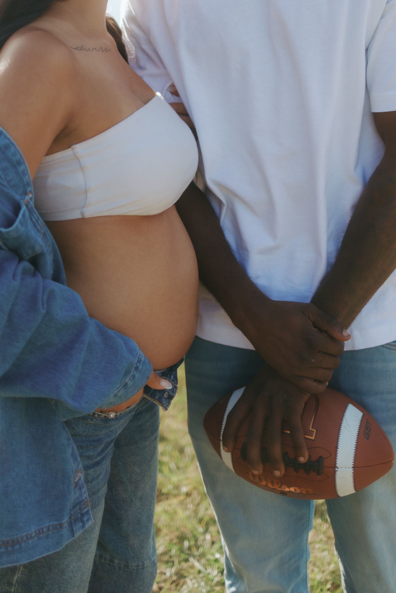 Pregnant woman in white crop top and denim jacket stands beside man holding football outdoors.
