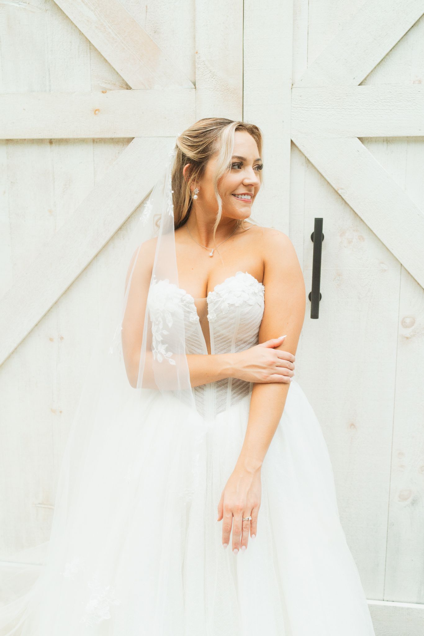 Smiling bride in strapless lace wedding gown and veil poses before rustic white barn doors.