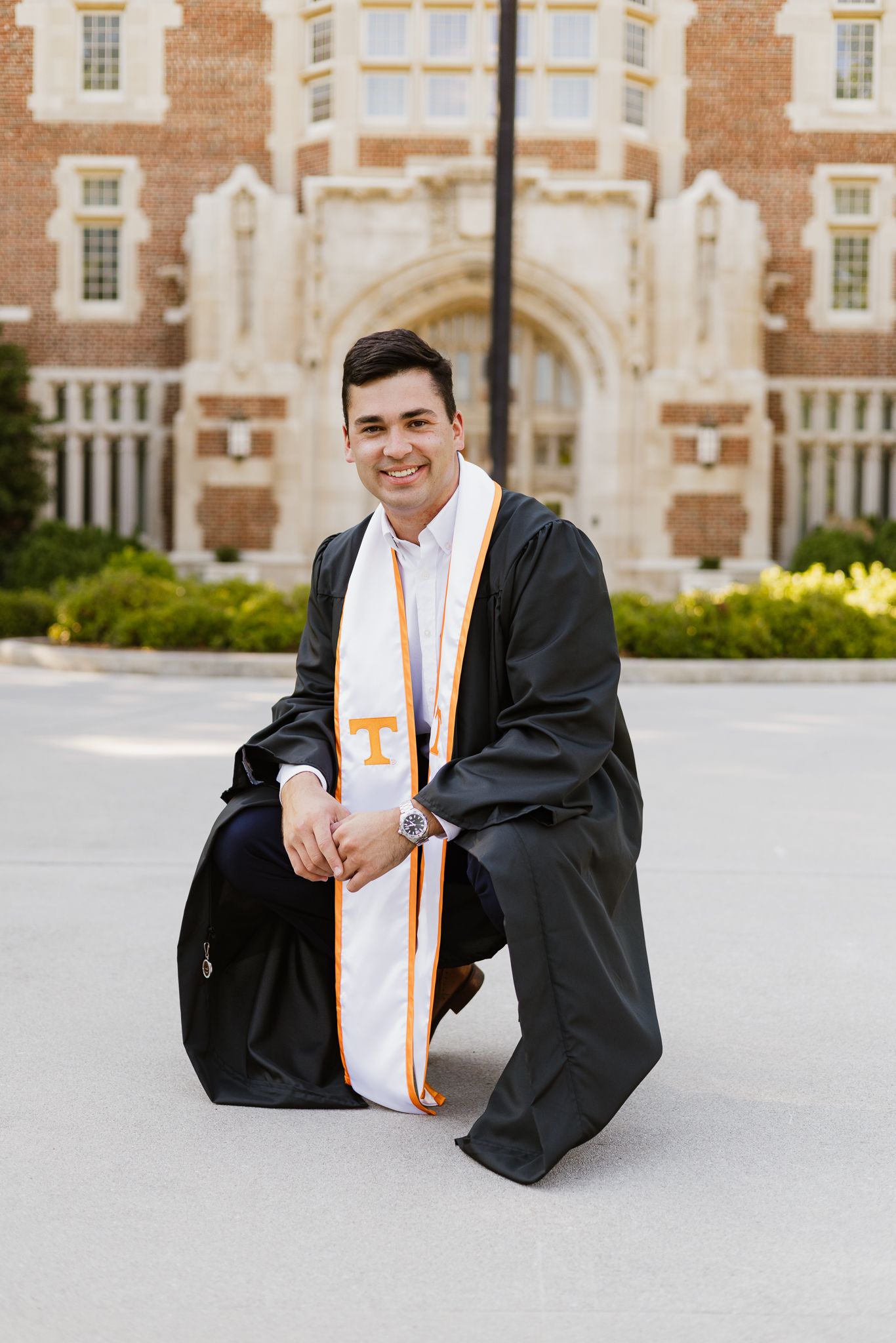 A University of Tennessee graduate kneels proudly in cap and gown with orange and white stole on campus.