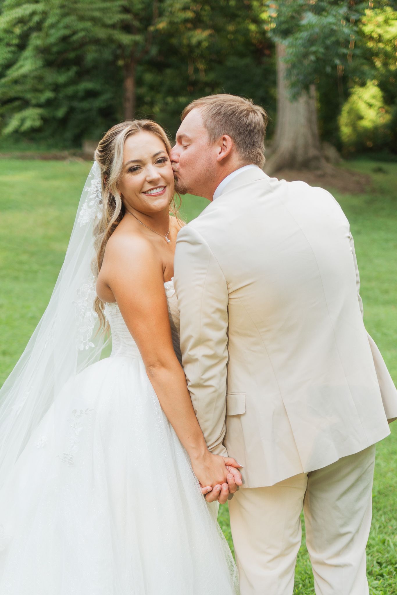 Groom in tan suit kisses bride on cheek as she smiles, holding hands in a garden setting.