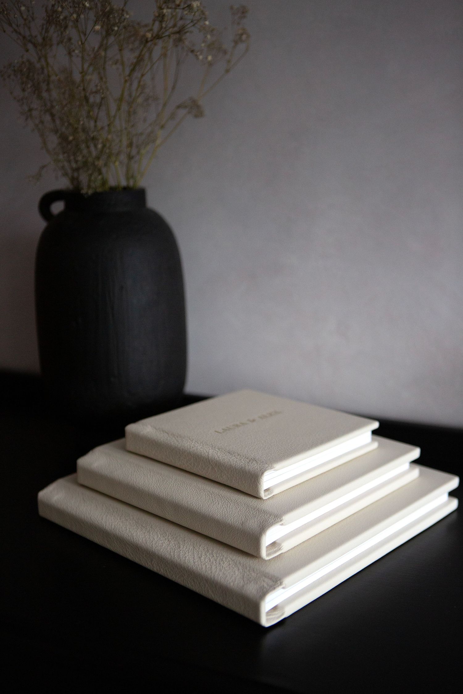 Stack of white wedding albums arranged by size next to a black vase with dried flowers on dark surface.