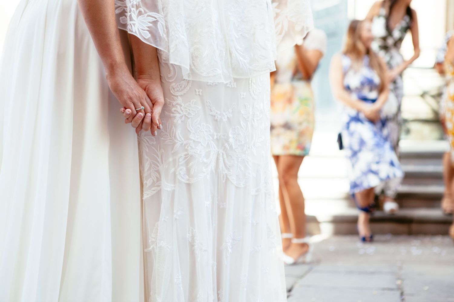Two brides hold hands against  a close-up detail of their wedding dresses with guests in floral outfits blurred in background on steps.