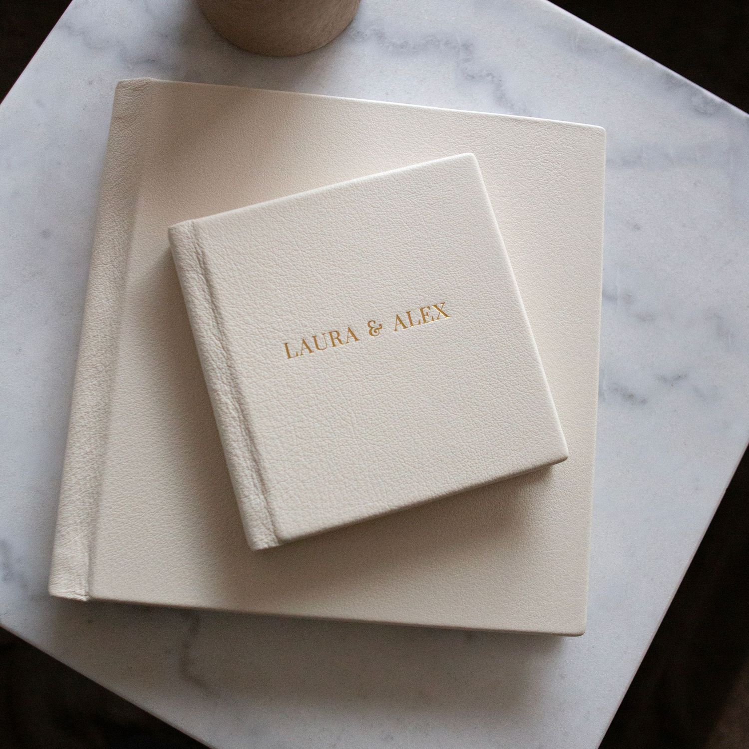 Two elegant white leather-bound wedding albums with gold lettering arranged on marble surface.