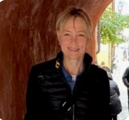 Smiling blonde woman in a black jacket standing outdoors under a rustic archway.