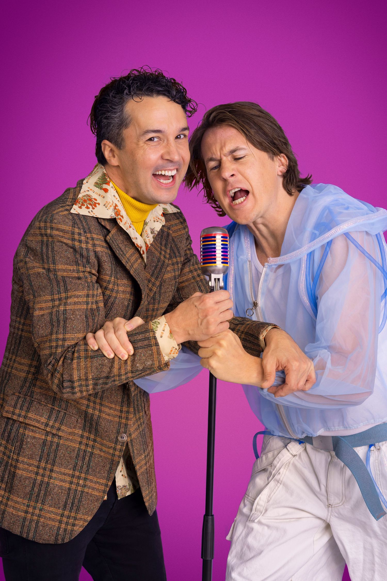 Two performers energetically singing into a vintage microphone against a purple background.