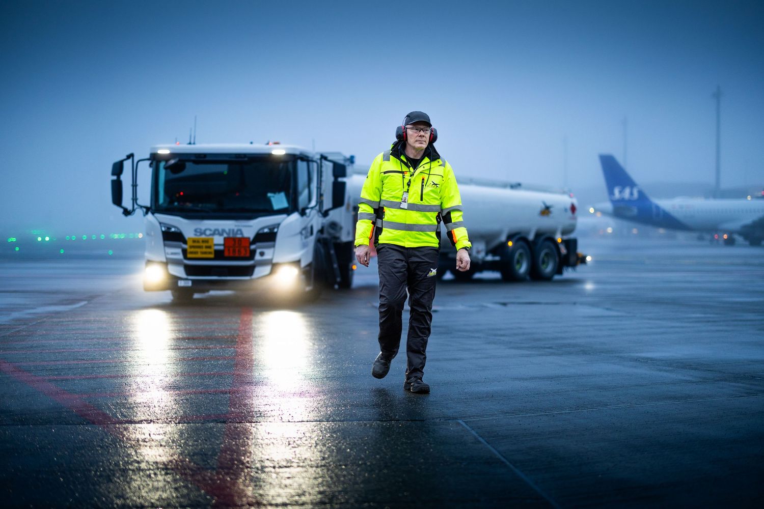 Airport ground crew worker in high-visibility jacket walks on wet tarmac at dusk.