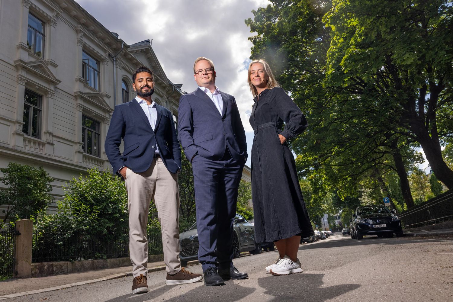 Three business professionals posing on street in front of historic building surrounded by trees.