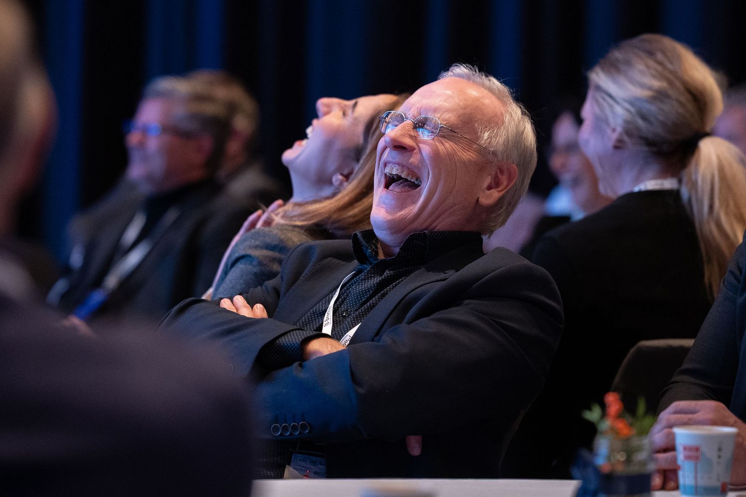Man laughs heartily with arms crossed while seated at a conference event with other attendees.