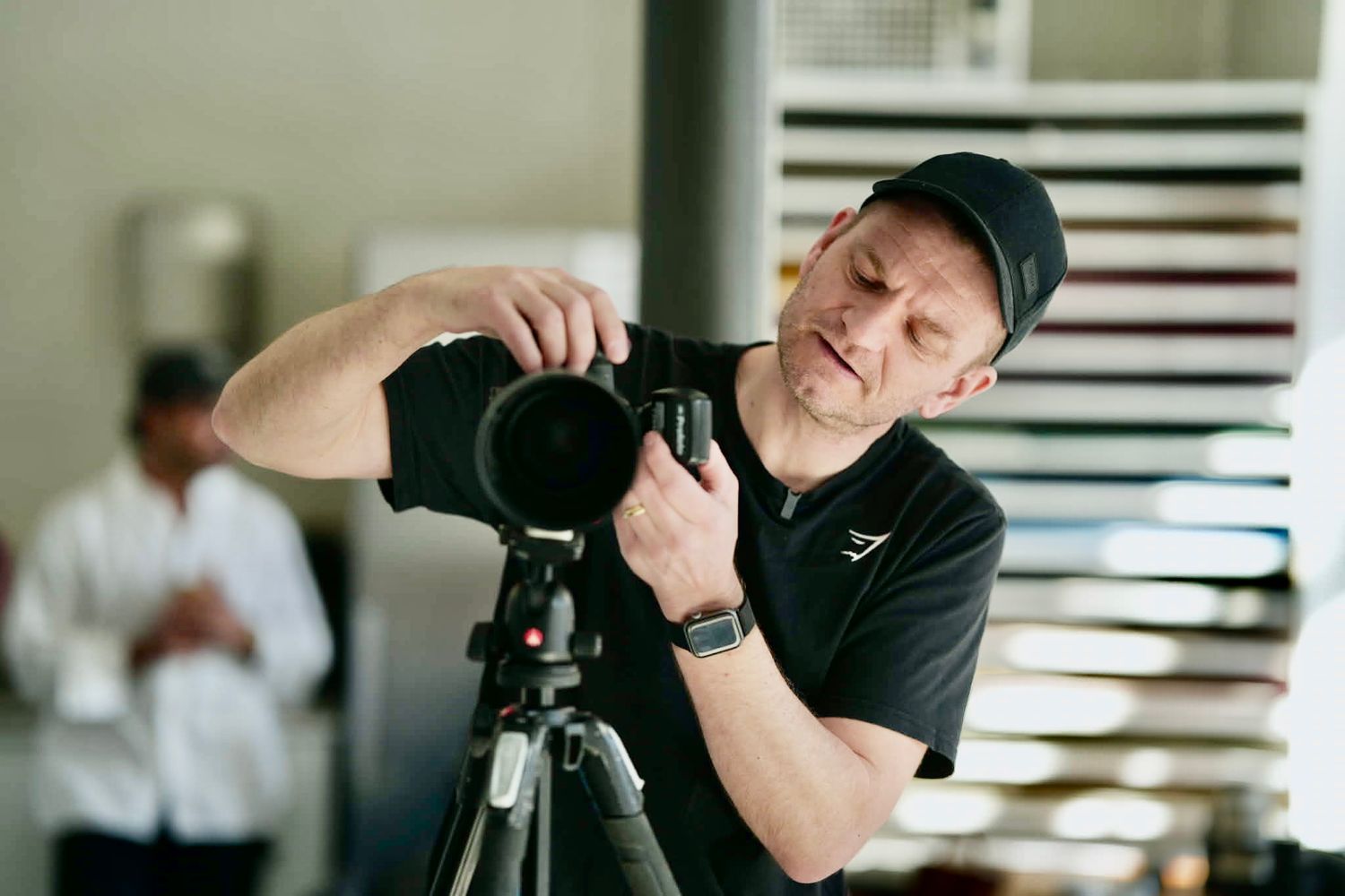 Man in a black cap and t-shirt adjusts a camera mounted on a tripod while a person in a white shirt stands blurred in the background.