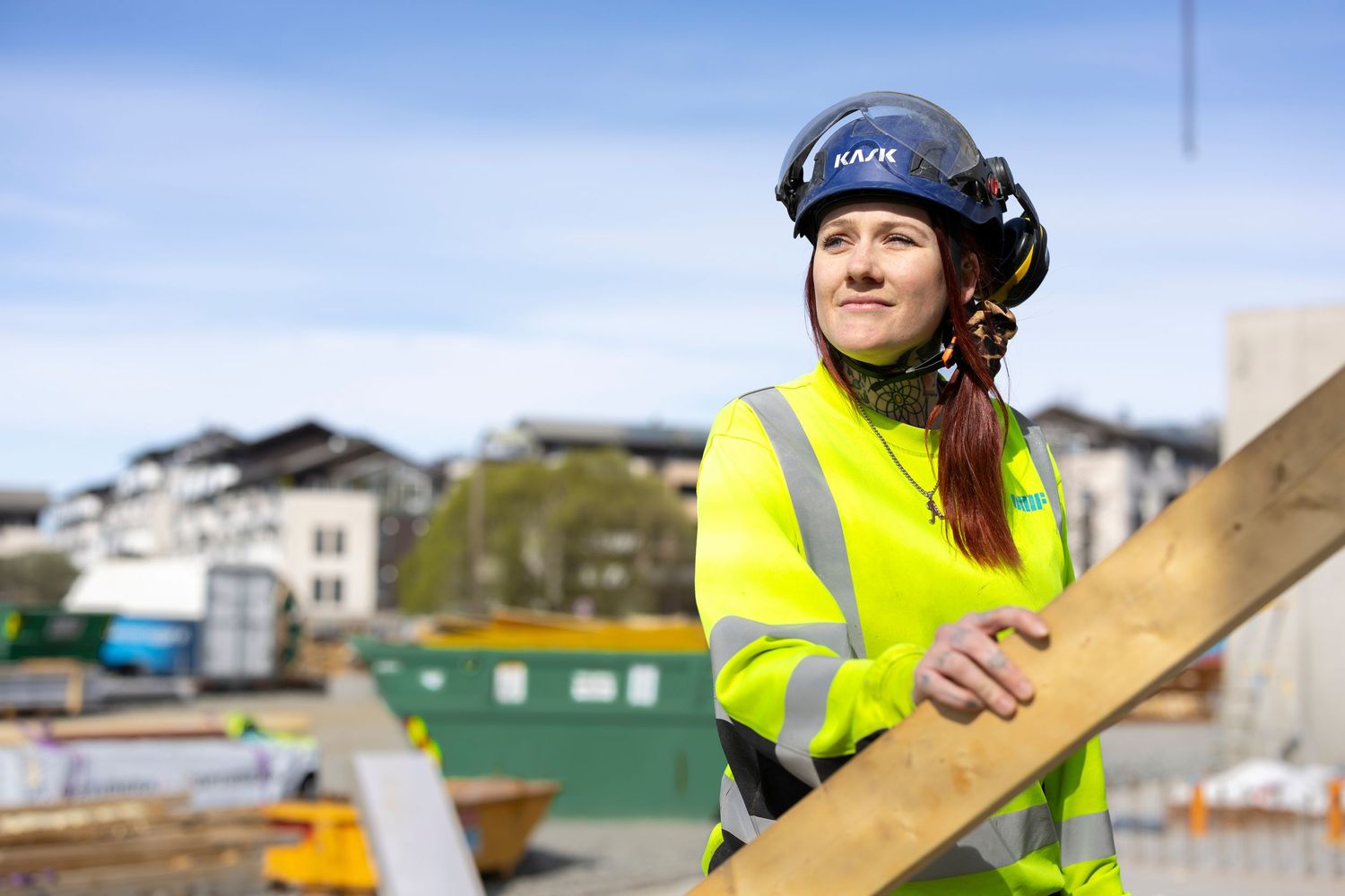 Female construction worker in yellow hi-vis jacket and blue KASK helmet holding timber at a building site.