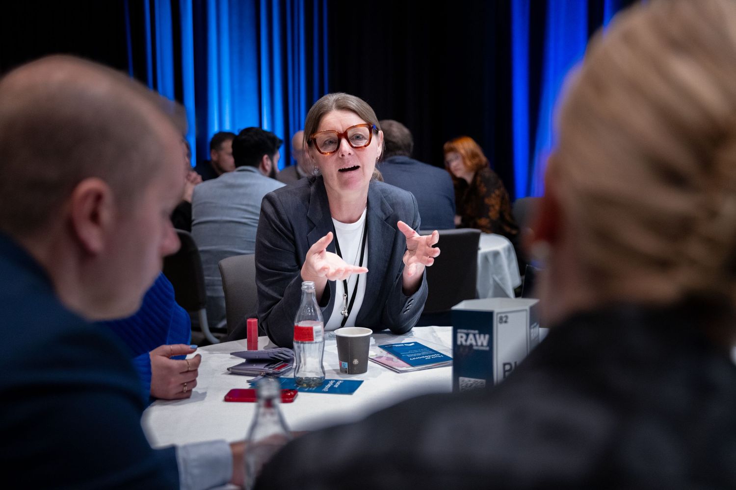 Woman with glasses gestures expressively during a discussion at a Nordic Forum RAW Materials conference table.