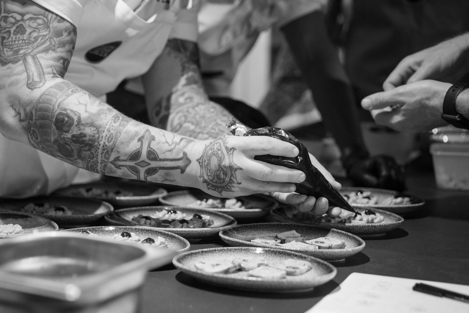 Tattooed chef's hands using scissors to garnish multiple small plated dishes at a culinary event.