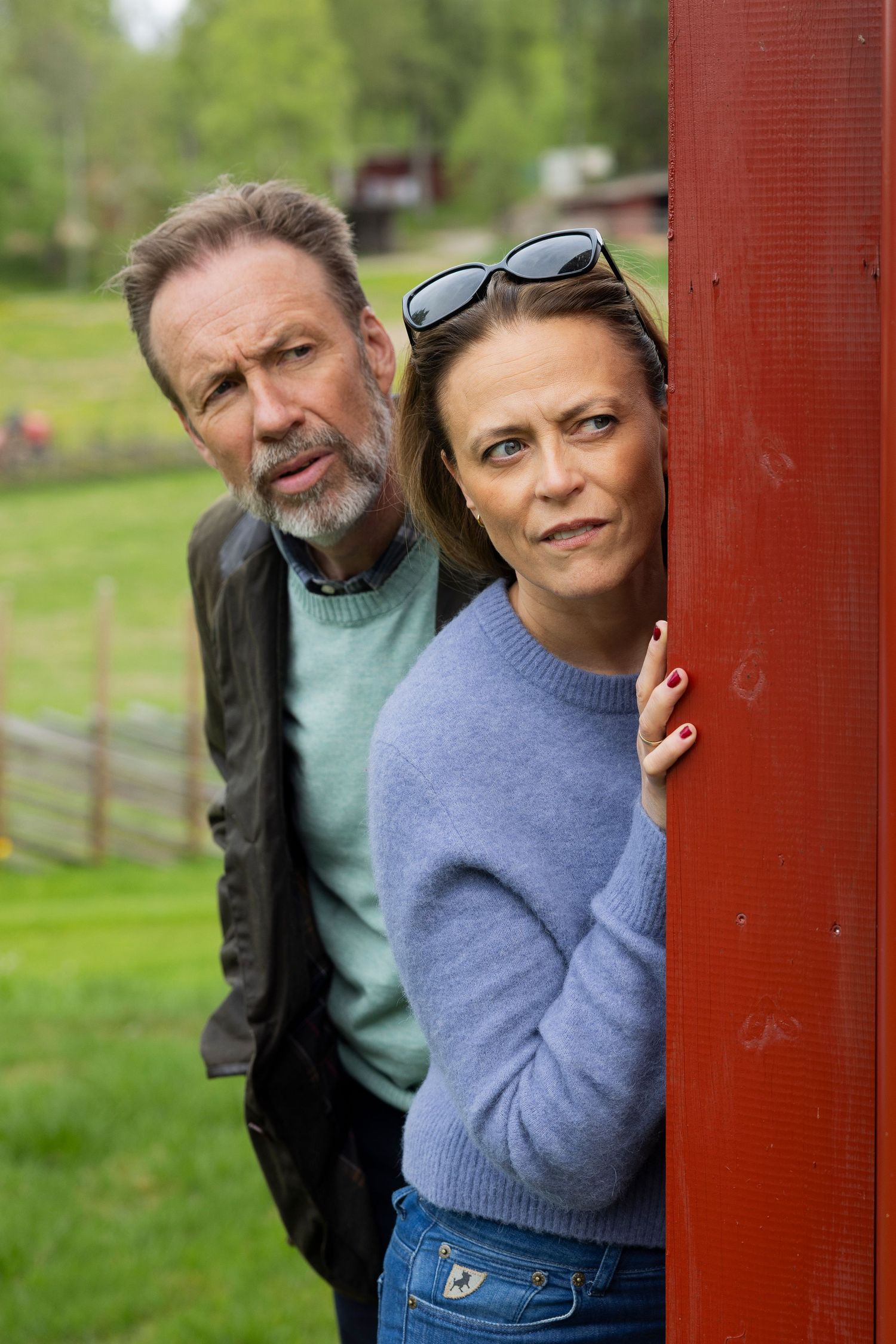 Man and woman with serious expressions peeking around a red wooden barn door outdoors.