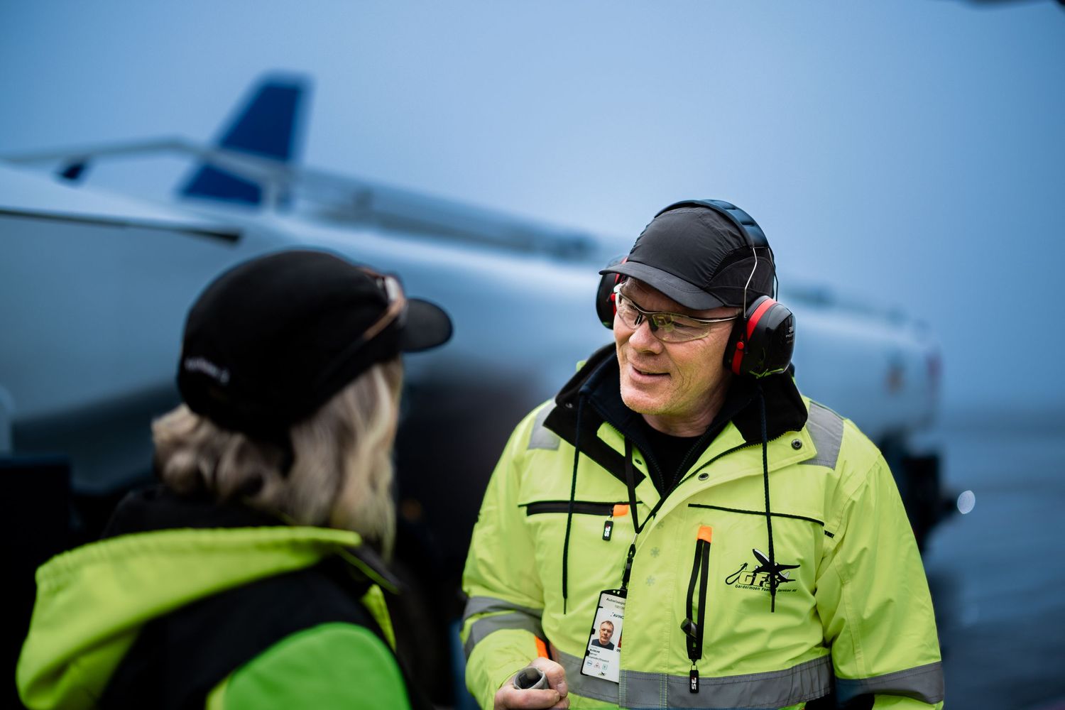 Two airport ground crew workers in yellow safety vests talk on tarmac near plane.