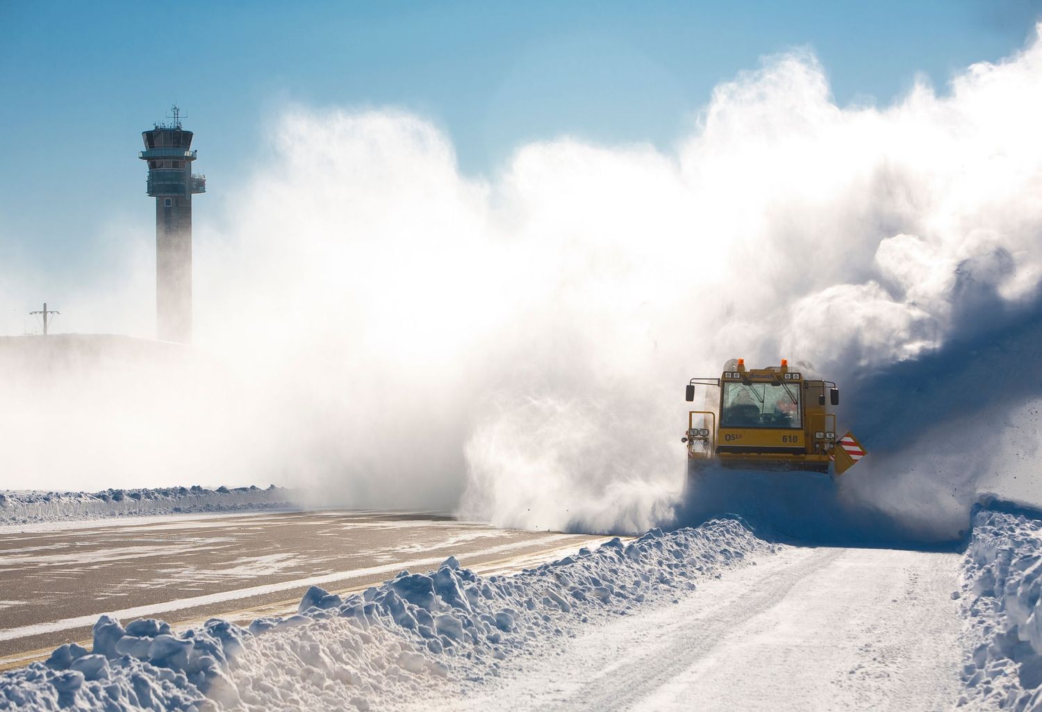 A yellow snowplow clears a runway in a massive cloud of snow at an airport on a bright winter day.