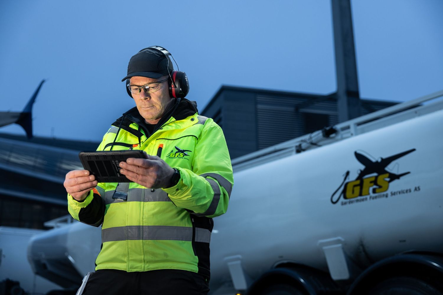 Airport ground crew worker in yellow safety jacket using tablet near aircraft at dusk.
