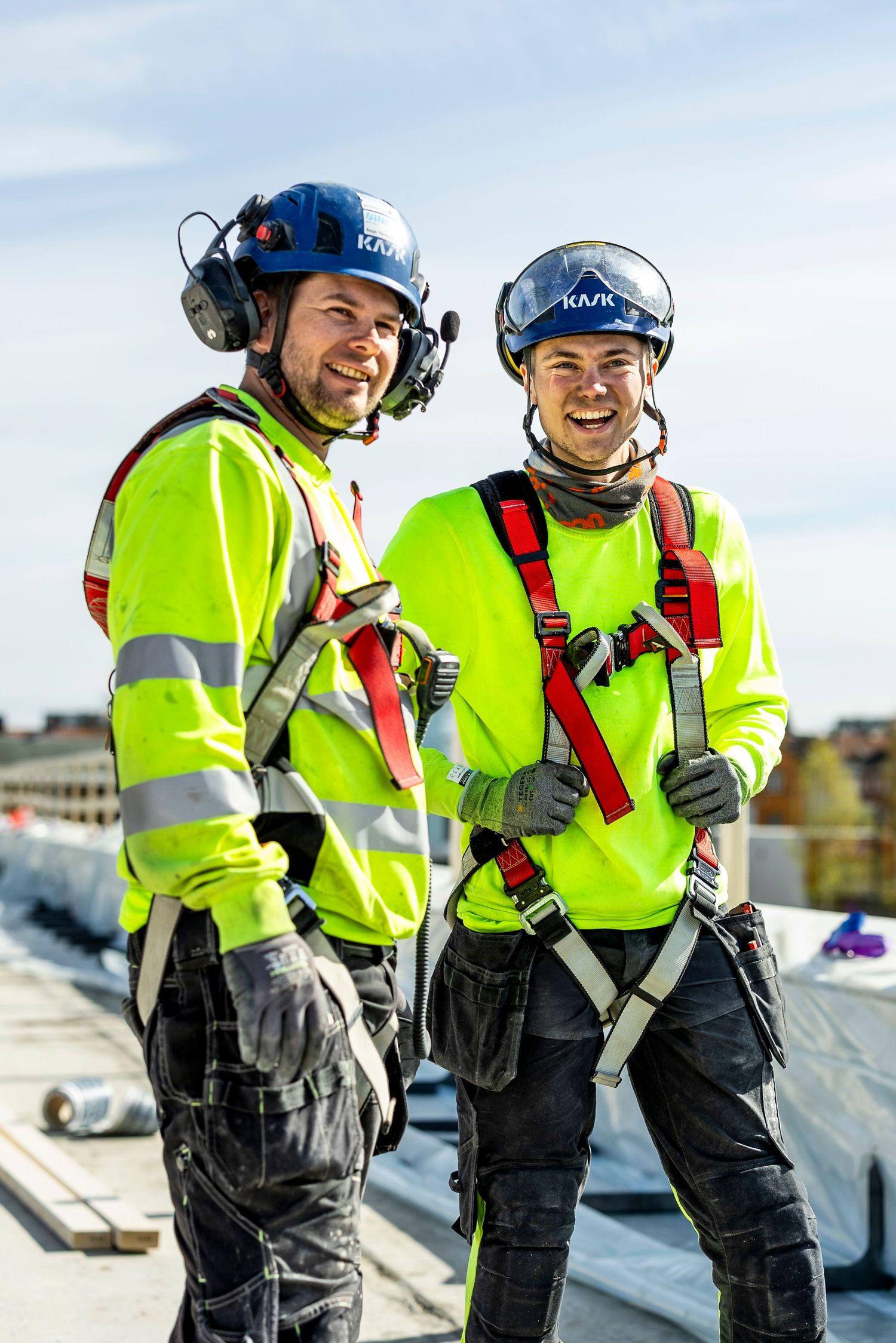 Two smiling construction workers in yellow hi-vis gear and safety helmets with harnesses pose on a rooftop construction site.