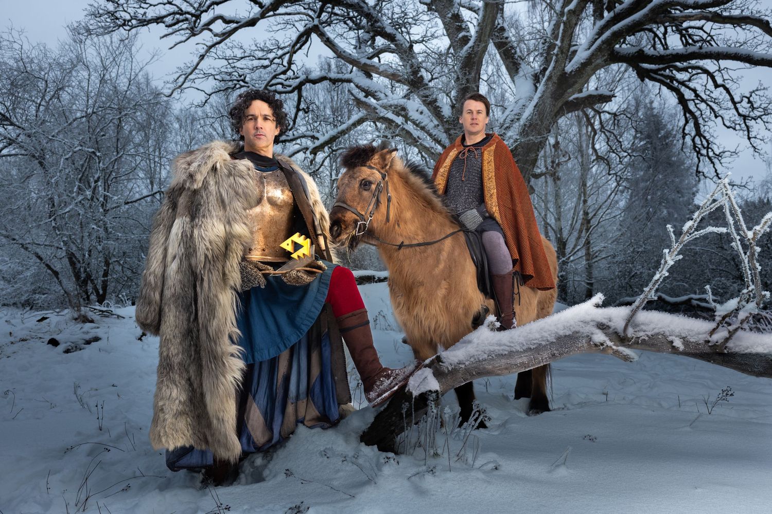 Two people in Viking costumes stand in a snowy winter forest landscape with a horse.