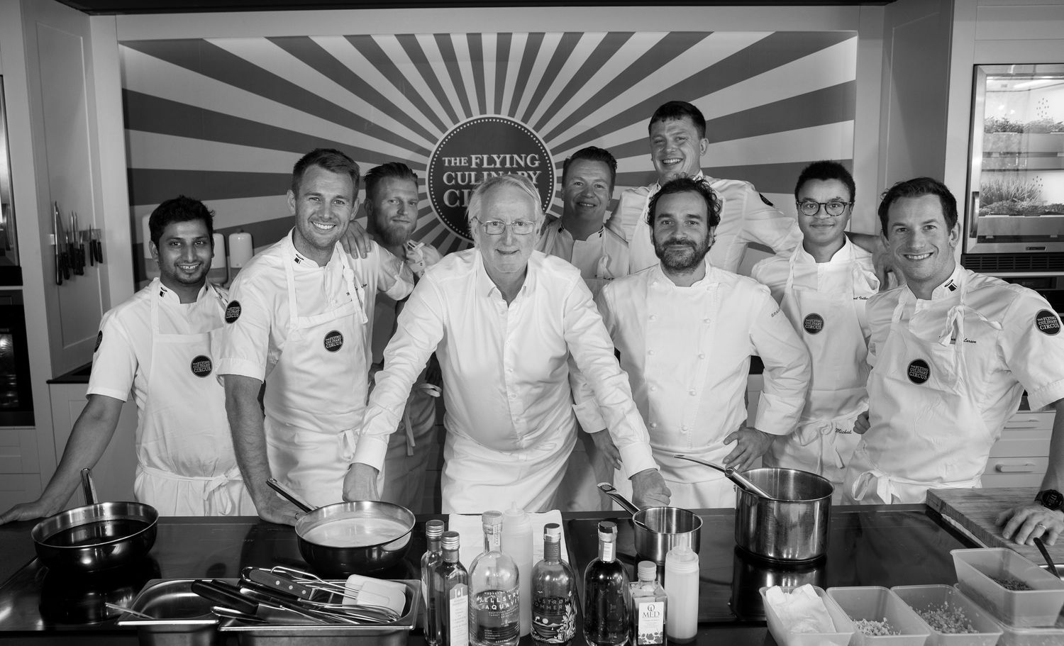 Group of chefs in white uniforms pose together at The Flying Culinary Circus kitchen event.