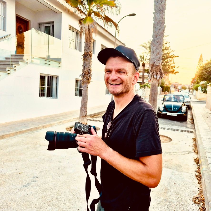 Smiling man holding a professional camera on a sunny street with palm trees and white buildings in background.