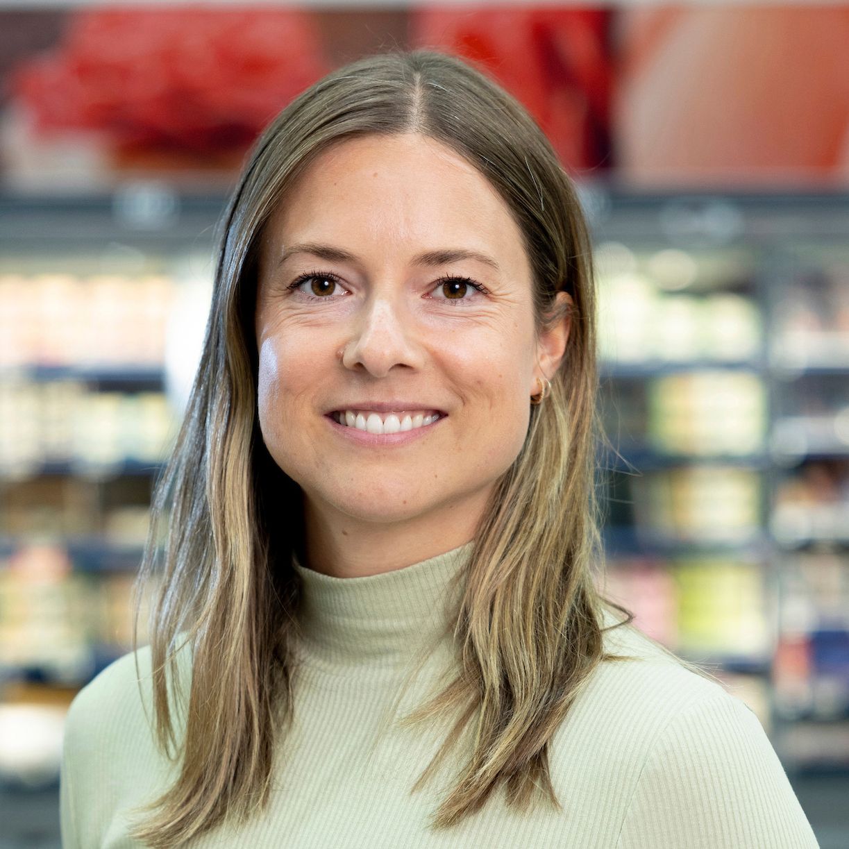 Smiling woman with long blonde hair wearing a light green turtleneck sweater standing in a grocery store.