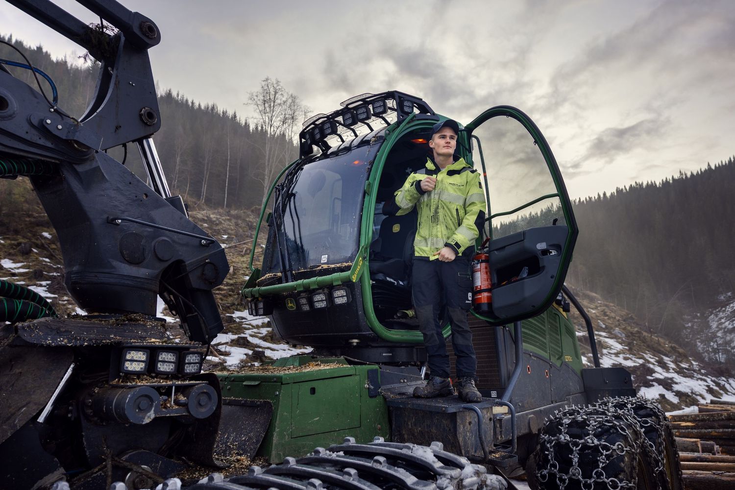 Forestry worker standing on a green harvester machine in a snowy forest.