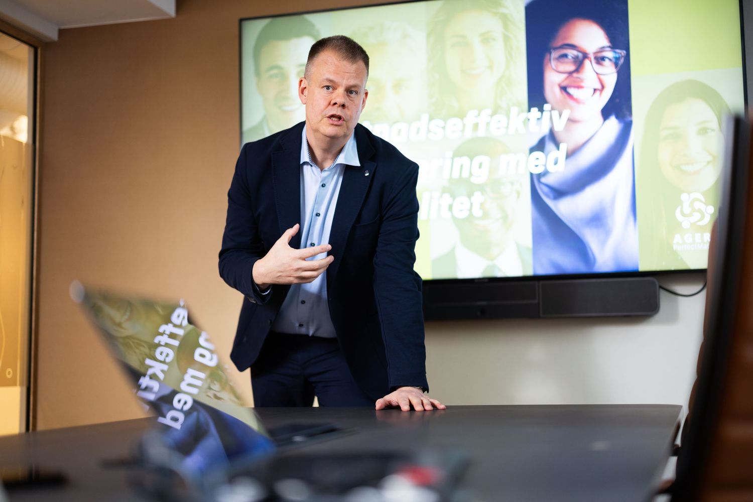 A man in a navy blazer presents in front of a screen displaying faces and text in a modern office setting.