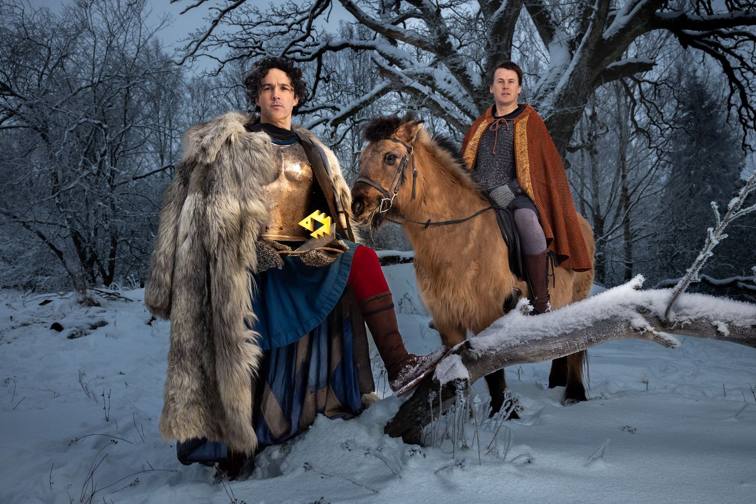 Two men in medieval costumes with a horse in a snowy winter forest setting.