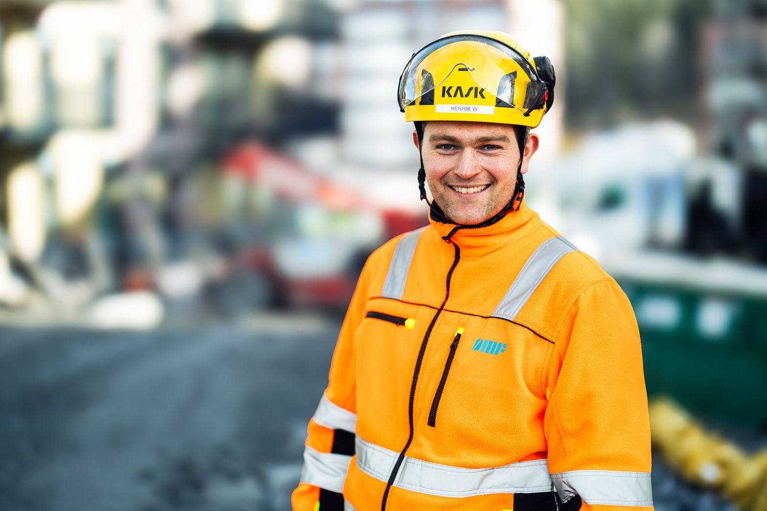 Smiling construction worker in orange hi-vis jacket and yellow KASK helmet at an outdoor building site.